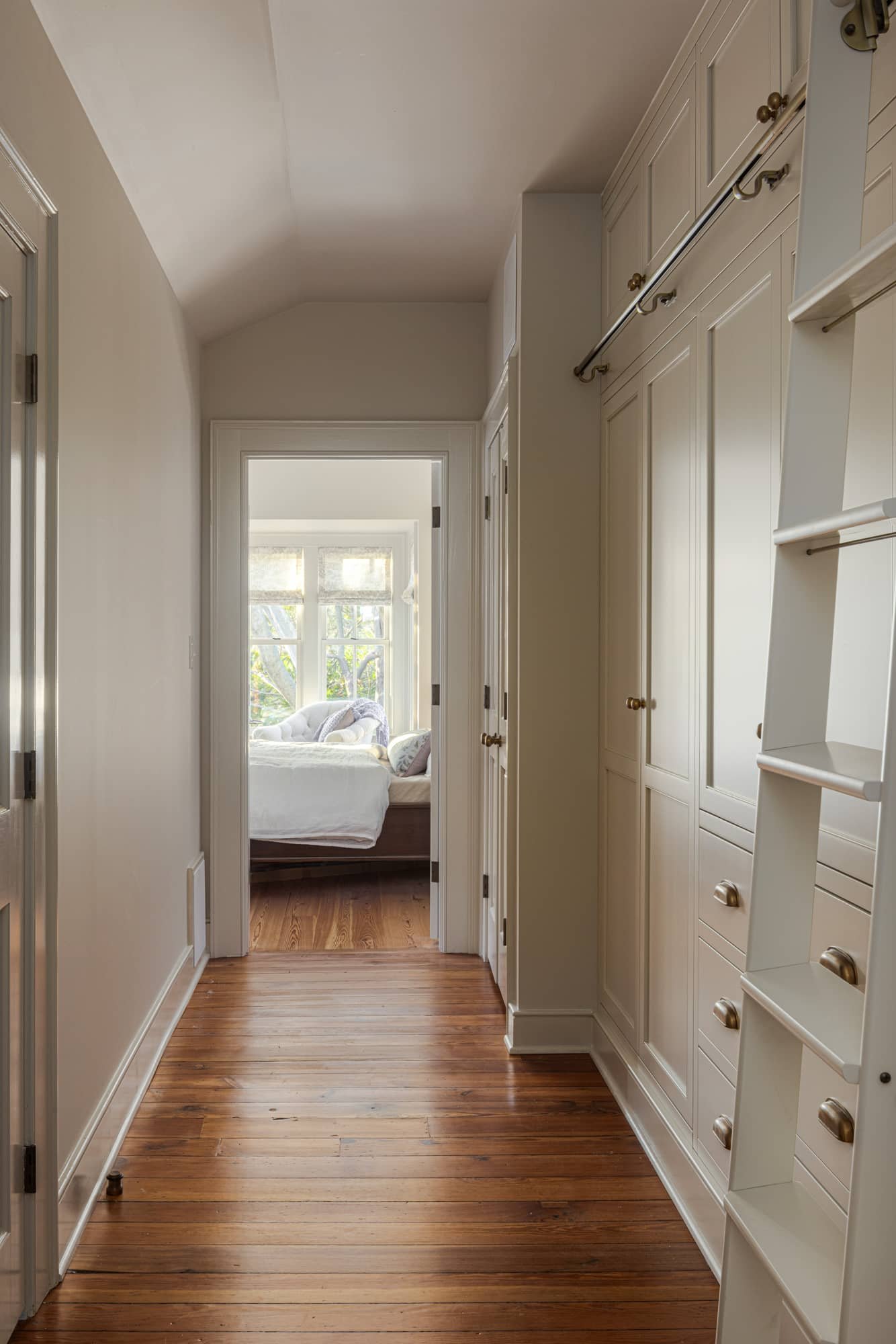 A hallway with white walls, wooden flooring, and built-in white cabinetry, leading to a sunlit bedroom with large windows and a bed with white bedding.