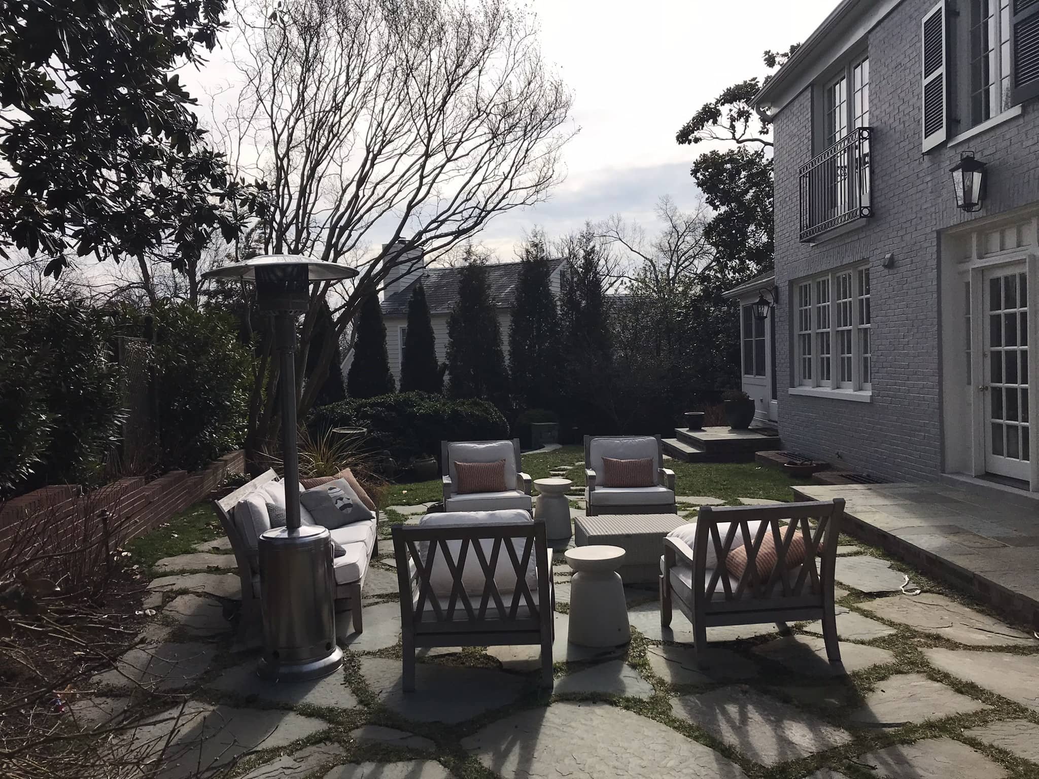 A backyard patio with outdoor seating including two chairs, a loveseat, and a small table, surrounded by plants and trees, with a large house on the right side and a patio heater on the left.