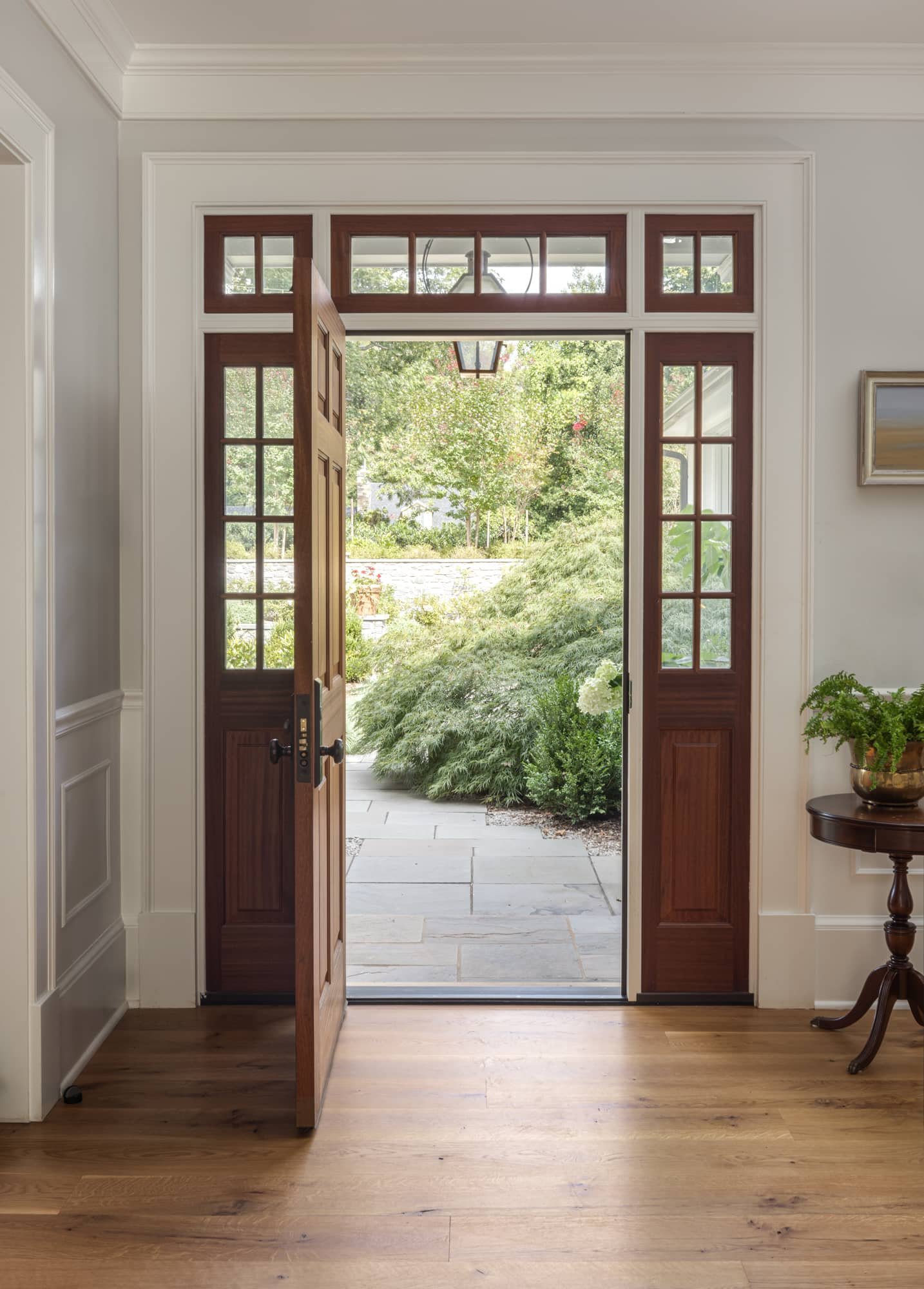 Open wooden front door leading outside to a garden with trees and bushes, bright sunlight, and stone patio, interior with hardwood floors and white walls.