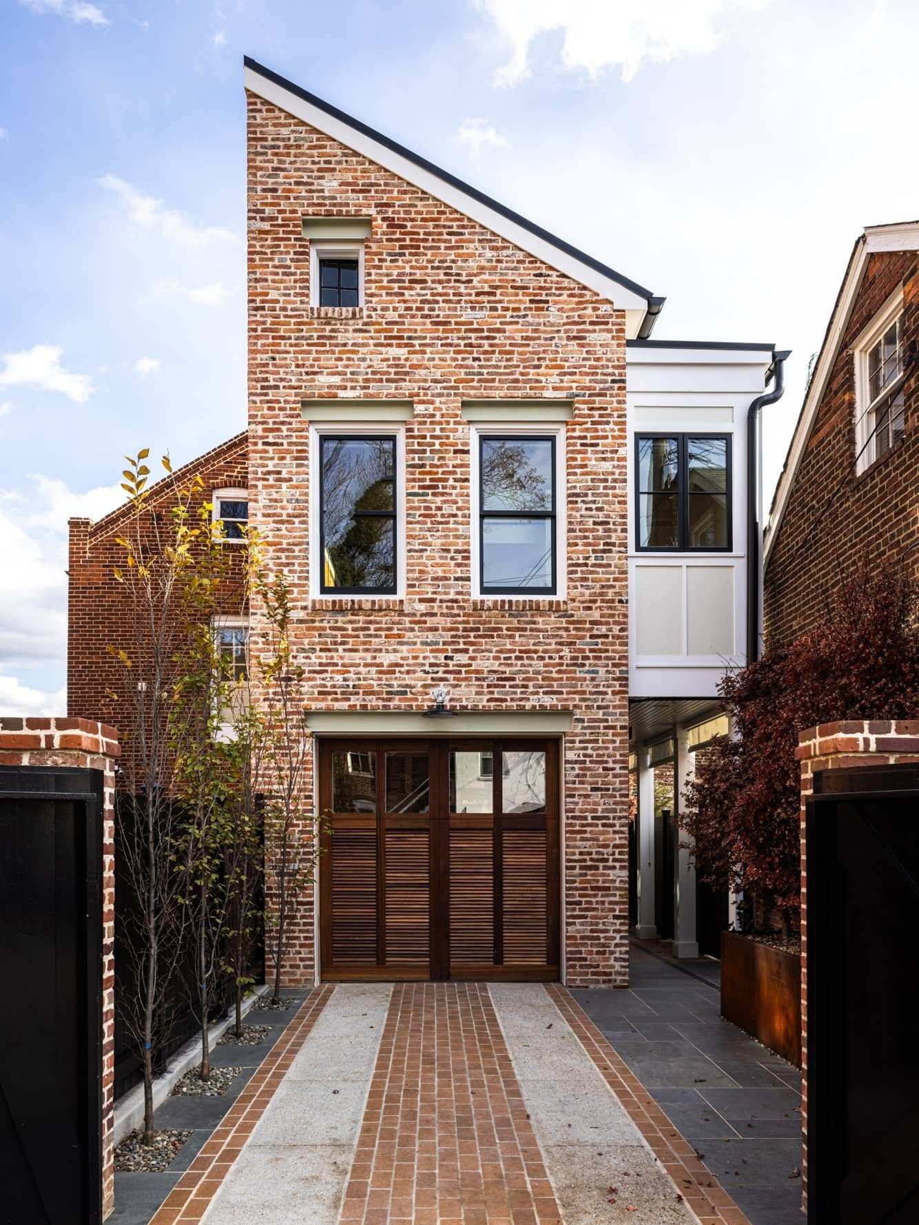 Modern brick house with a sloped roof, two large windows on the second floor, and a garage door on the ground level. The driveway features brick and concrete patterns, flanked by small trees and black gates.