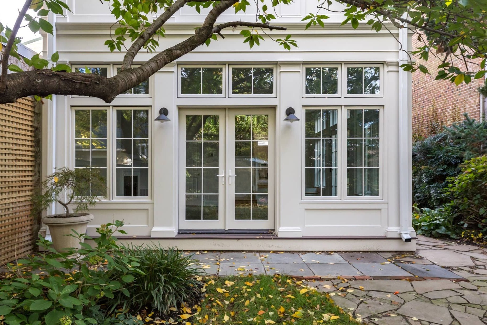 A white house with glass room and large windows and a glass double door, surrounded by greenery, trees, and a stone patio in the backyard.