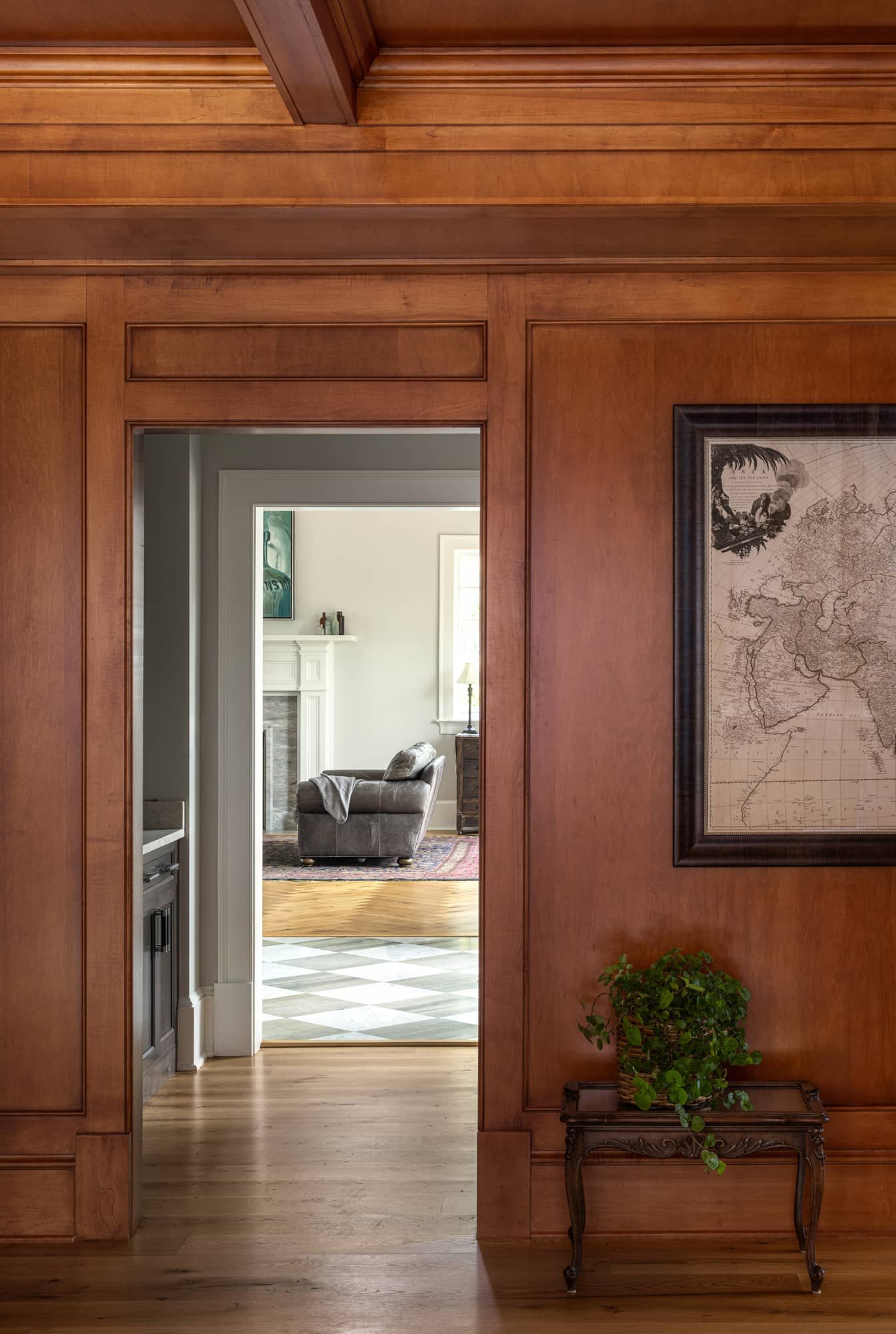 View through a doorway into a living room with a gray couch, fireplace, and window with a lamp. The foreground features a wooden wall with a framed map and a small table with a green potted plant.