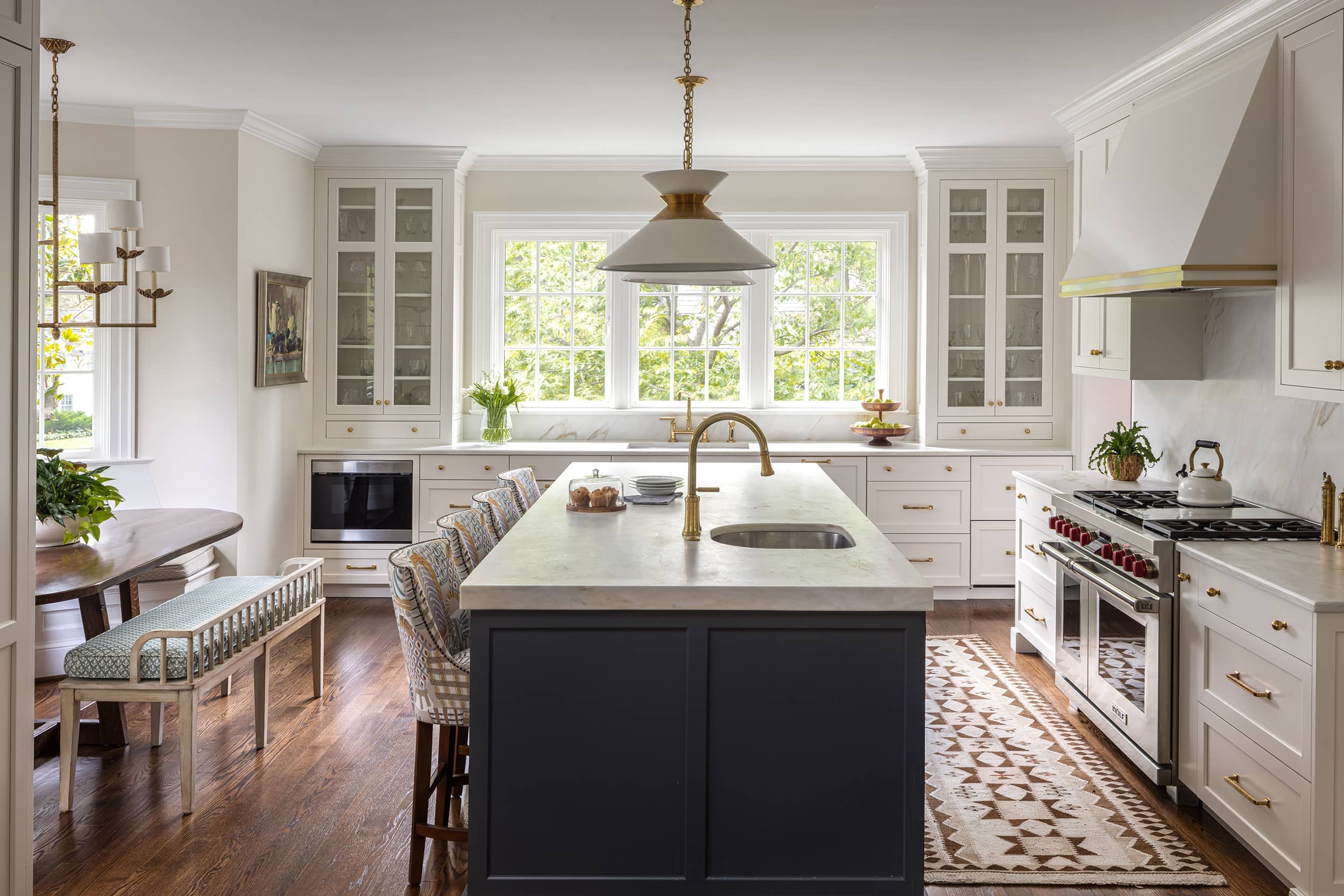 Bright, white kitchen with large window, island with a sink, and surrounding cabinets. Gold accents on fixtures and handles. Wooden flooring, patterned rug, and decorative items like potted plants and a cake stand.