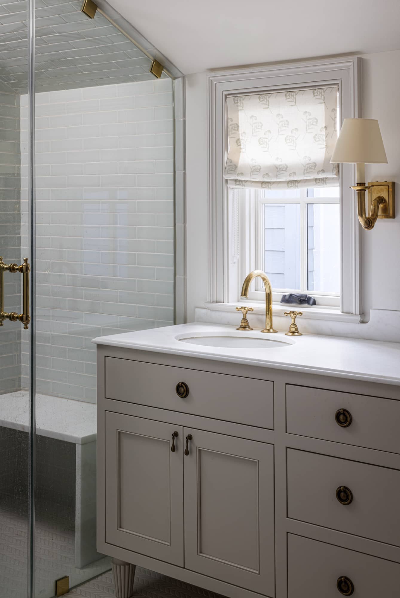 Bathroom with white vanity, gold fixtures, window with patterned curtain, and glass-enclosed shower with white subway tiles.