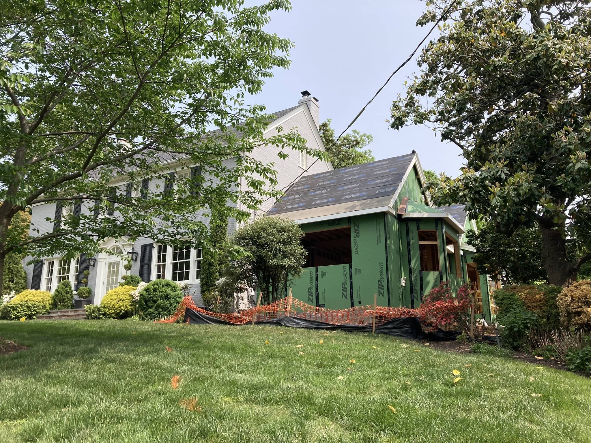 A house under construction with a green exterior wall, a partially installed roof, surrounded by trees and bushes, with a grassy lawn in the foreground.