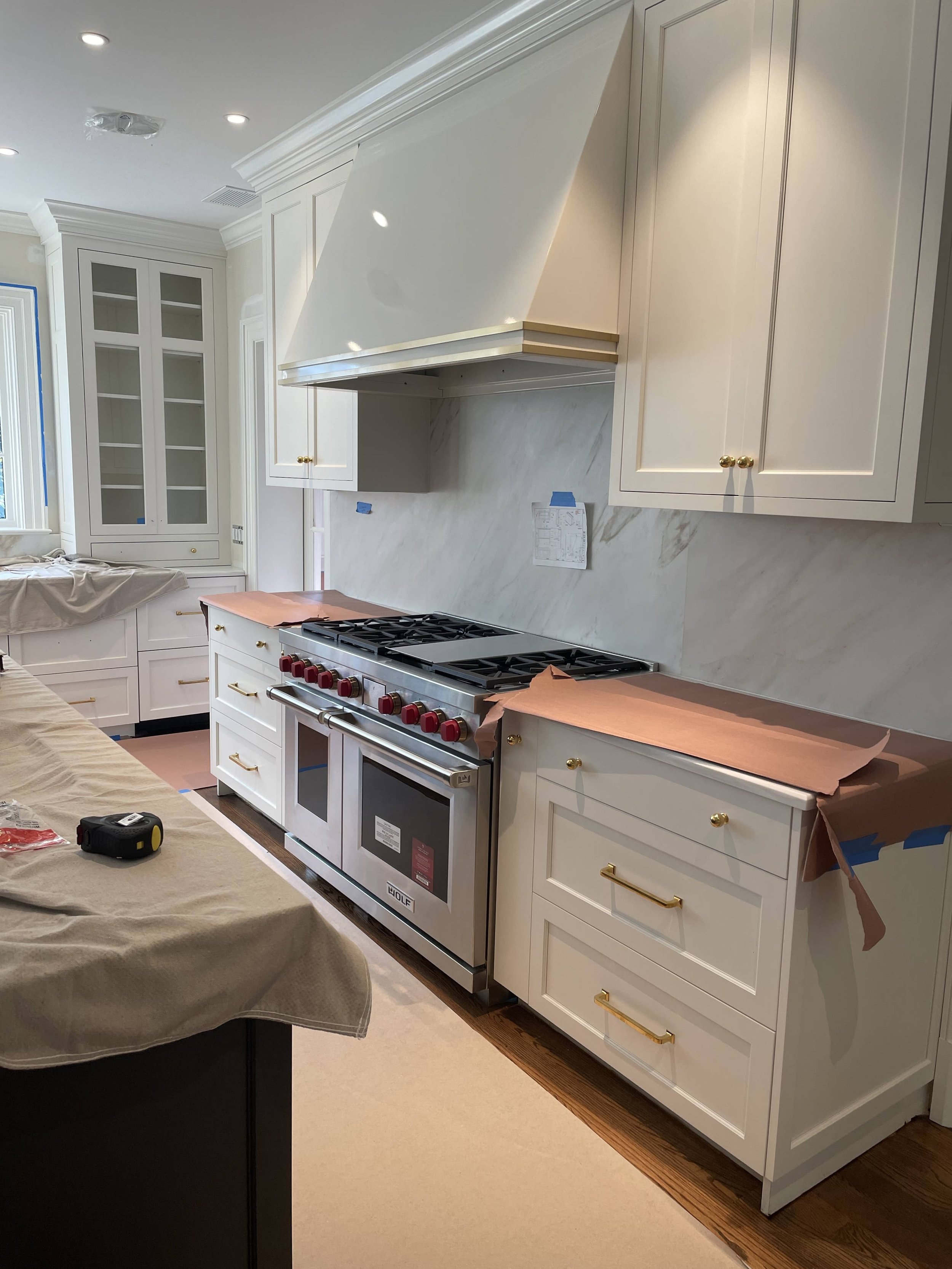 Kitchen with white cabinets and gold handles, a large stove with red knobs, a white marble backsplash, and some blue painter's tape on the wall. There is a cooking range cover in progress, and a workspace with a work tape measure.
