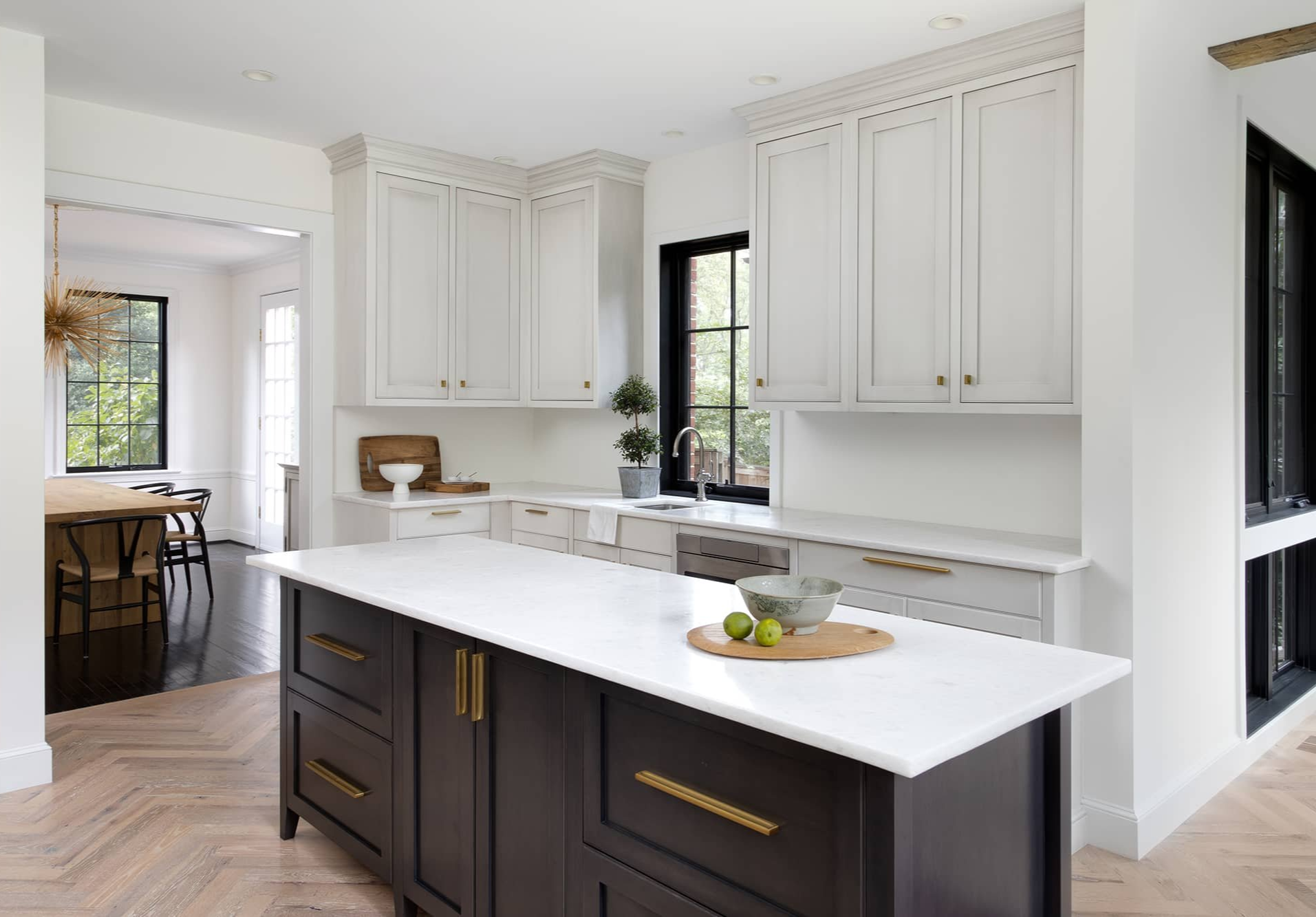 Modern kitchen with white cabinets, black window frames, and a black kitchen island with gold handles, with a fruit bowl and limes on the island. Adjacent dining area with a wooden table and black chairs.