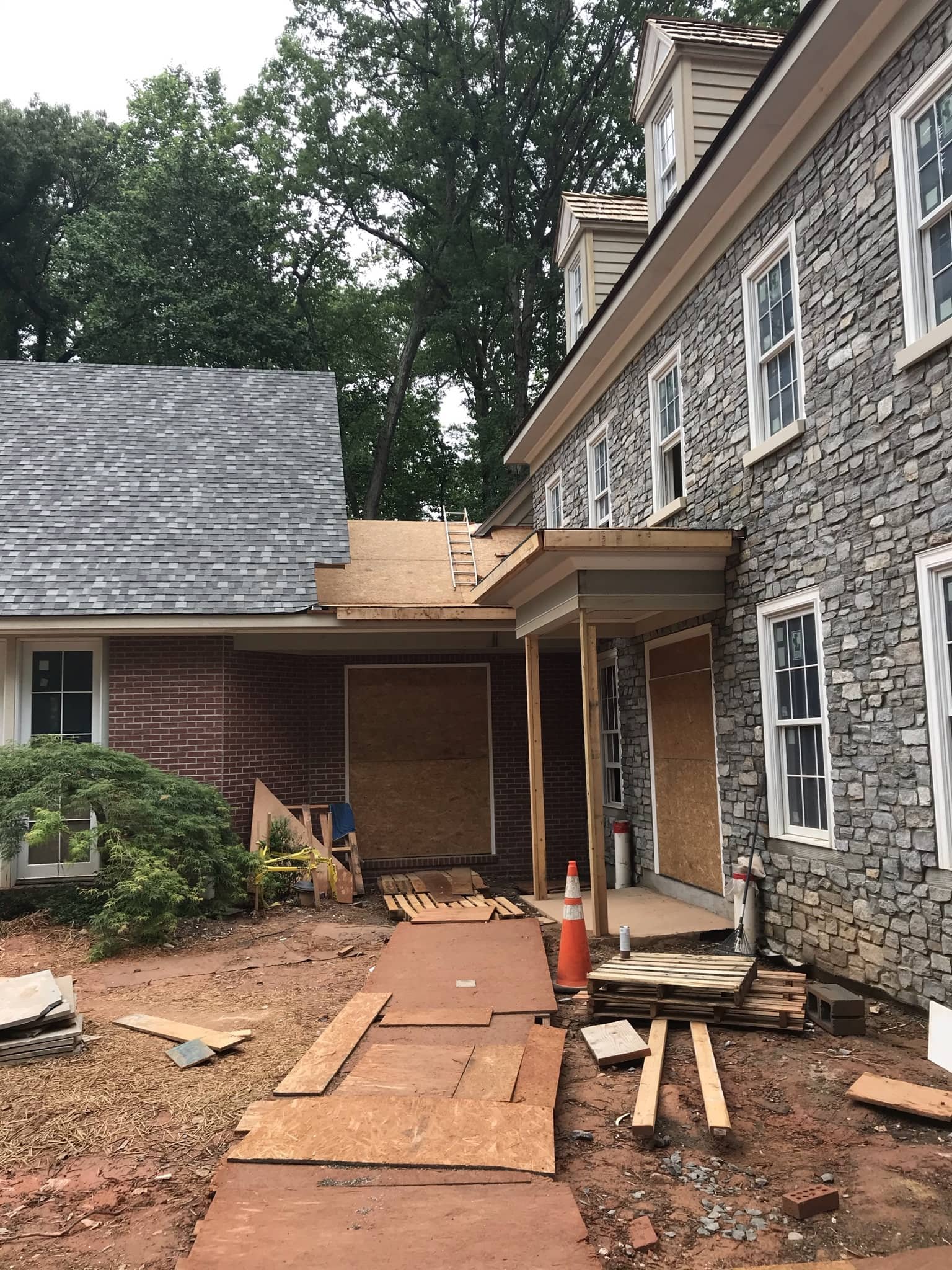 Construction site showing a partially built extension with plywood on the walls and roof, outdoor tools, and building materials around.