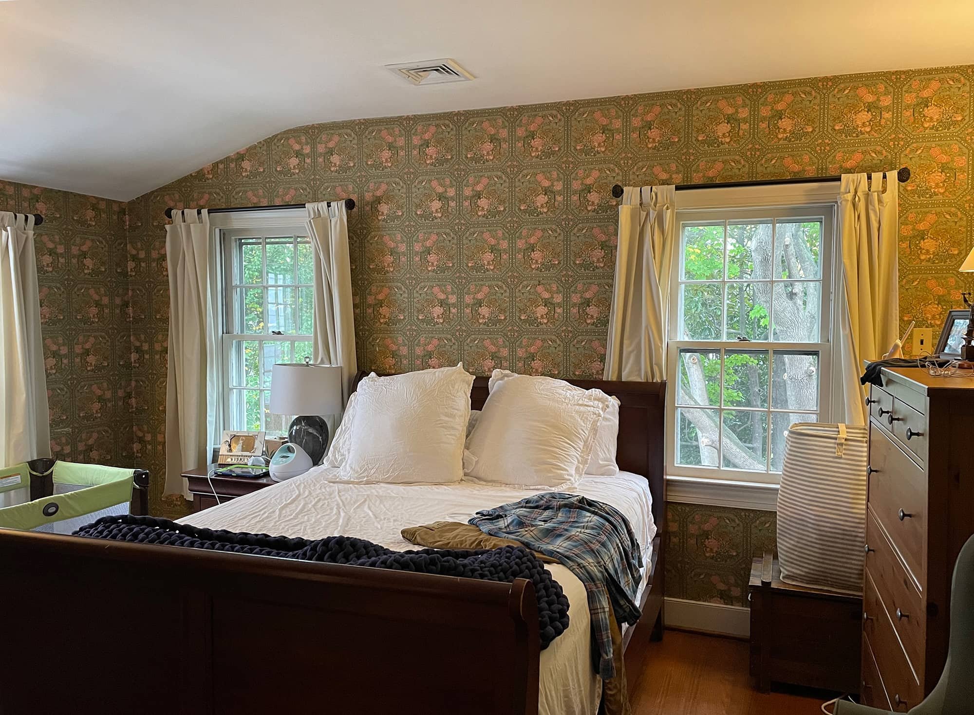 Comfortable bedroom with a wooden bed frame, white bedding, and pillows, two windows with cream curtains, a nightstand with a lamp and photo frame, a dresser with a lamp and items, a laundry basket, a partially visible green playpen, and a cozy, floral-patterned wallpaper.