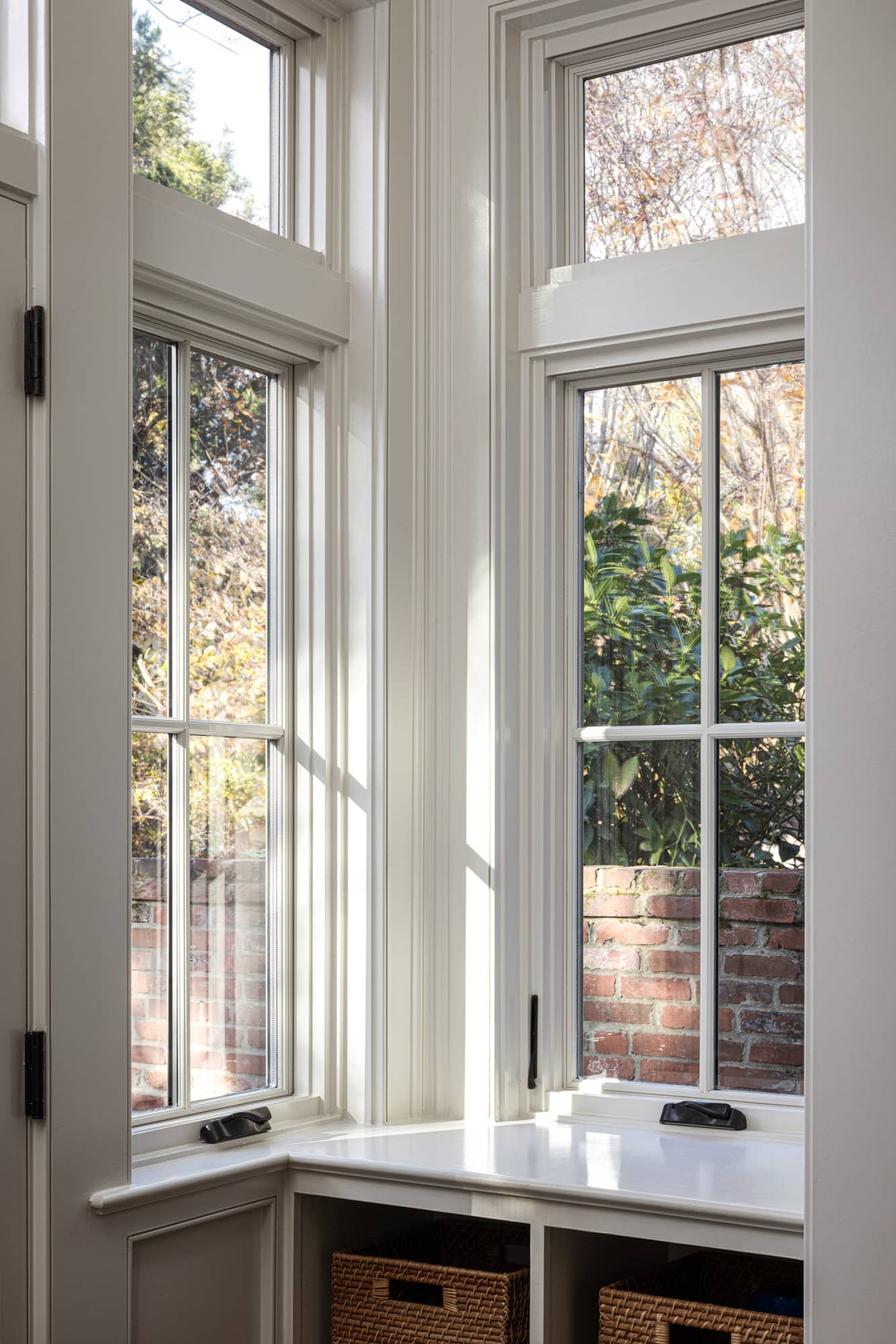 A corner of a room with three tall, double-pane windows framed in white, featuring latch handles at the bottom. Sunlight streams through, casting shadows on the window frames and the windowsill. Outside, there are trees and a brick wall visible.