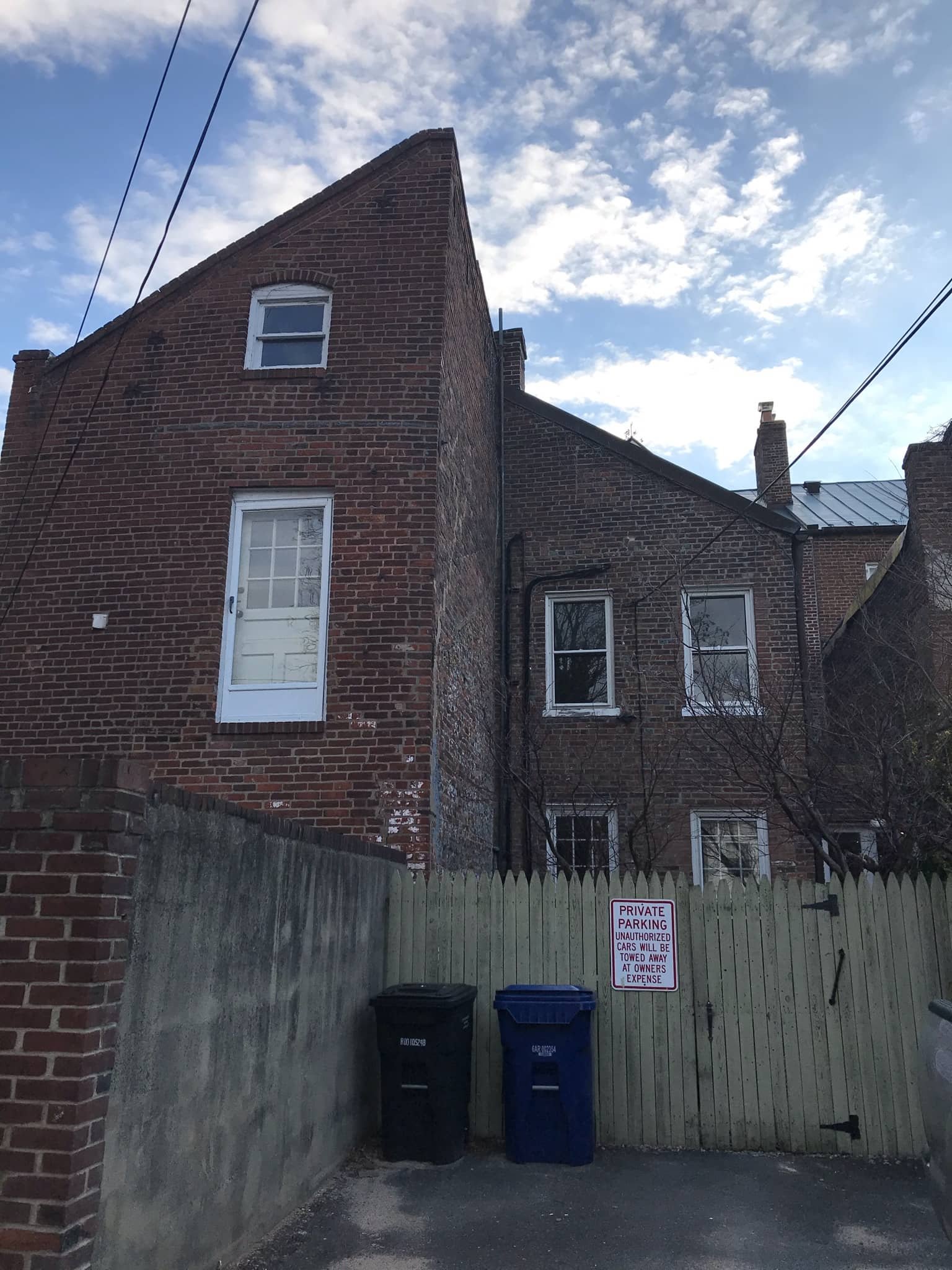 A brick building with multiple windows and a white door, a wooden fence with a sign that says 'Private Parking'. There are two garbage bins in front of the fence, one black and one blue. The sky is partly cloudy.
