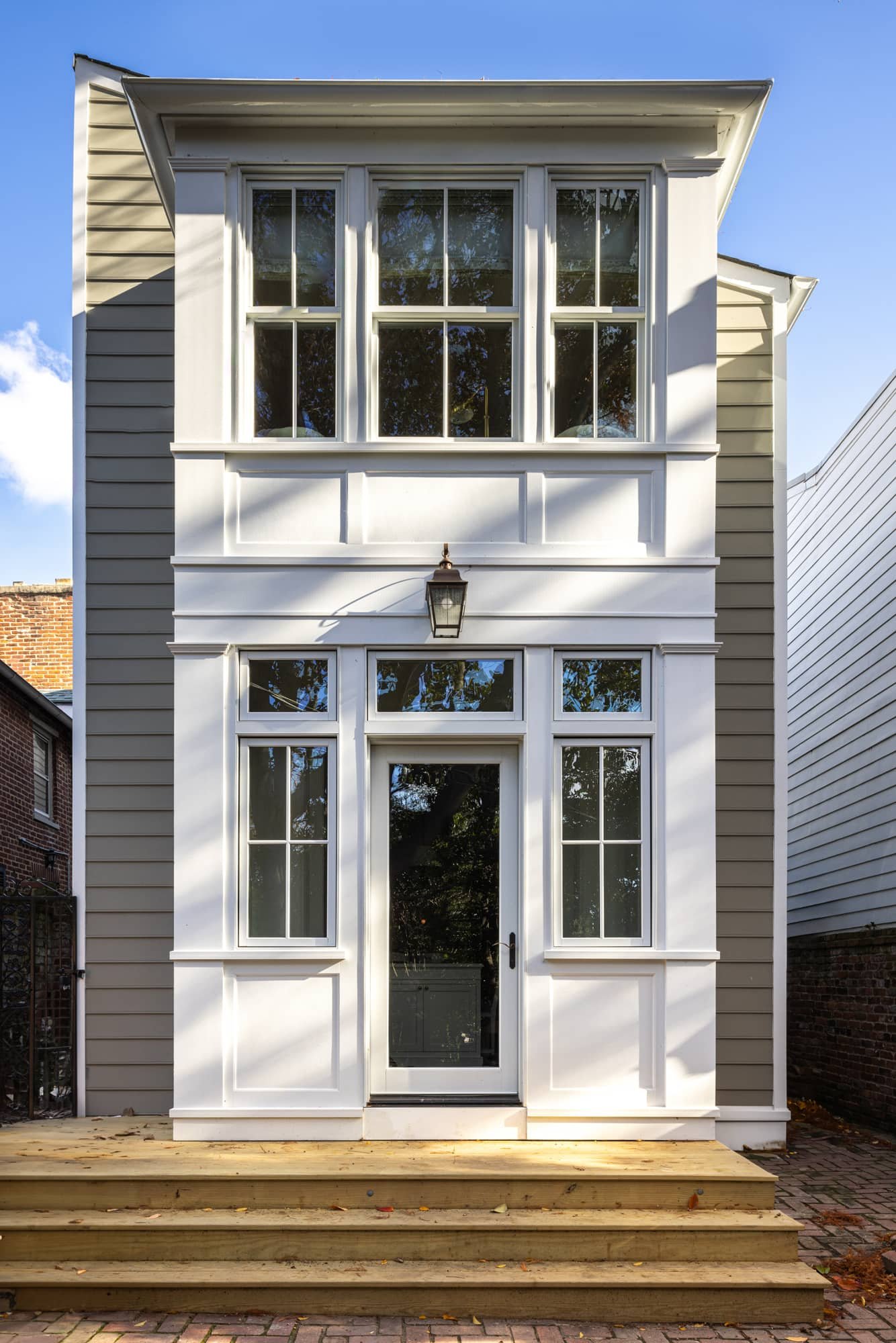 Front view of a modern three-story house with white siding, large windows, a black front door, a porch with wooden steps, and a brick and pavement pathway.