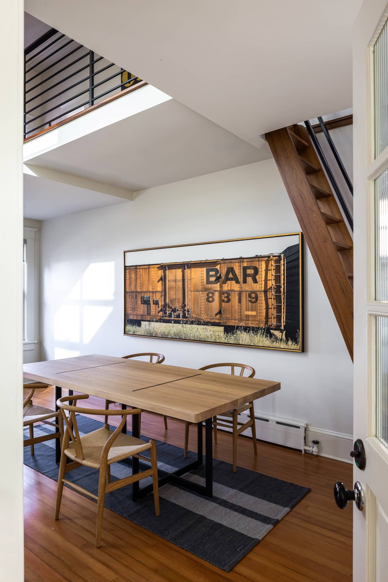 A dining room with a wooden table and six chairs, a large framed train car picture on the wall, a window letting in natural light, a staircase with wooden steps and black railing, and hardwood floors with a black and gray striped rug.