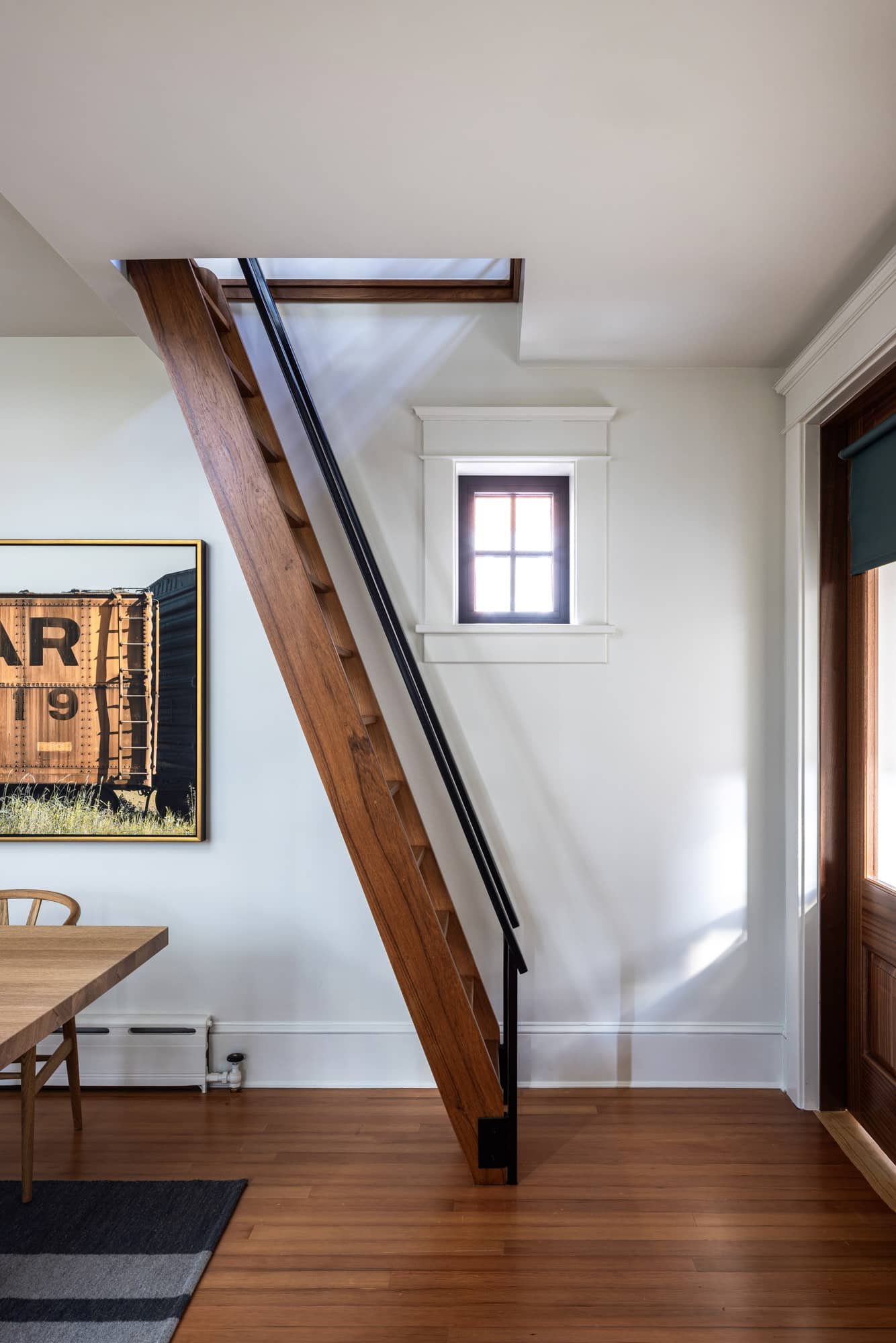 Interior view of a room with a wooden staircase, small window, and minimal furniture, with sunlight streaming through the window.