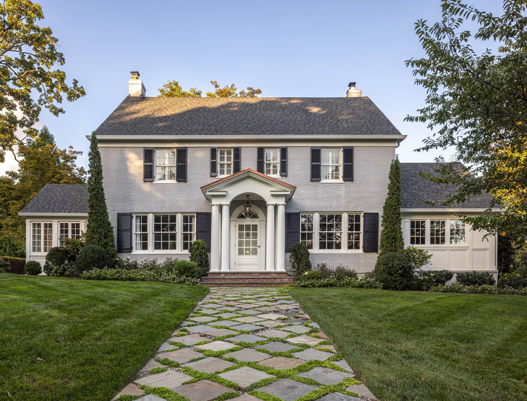 A large two-story historic colonial house with light brick exterior, black shutters, a brick pathway leading to the front door, and surrounded by lush green lawn and trees.