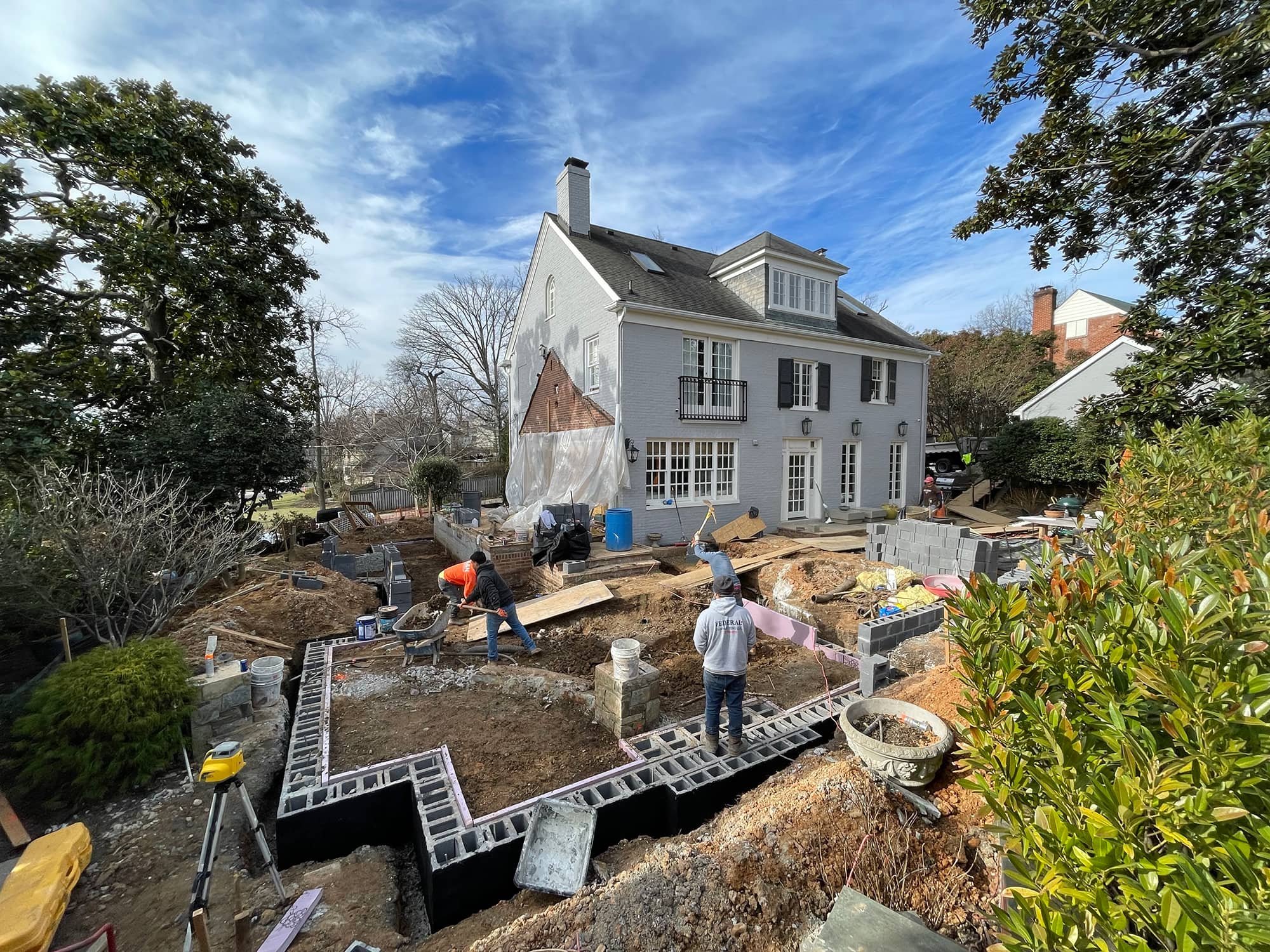 Construction workers building a backyard patio area in front of a large white house on a clear, sunny day.