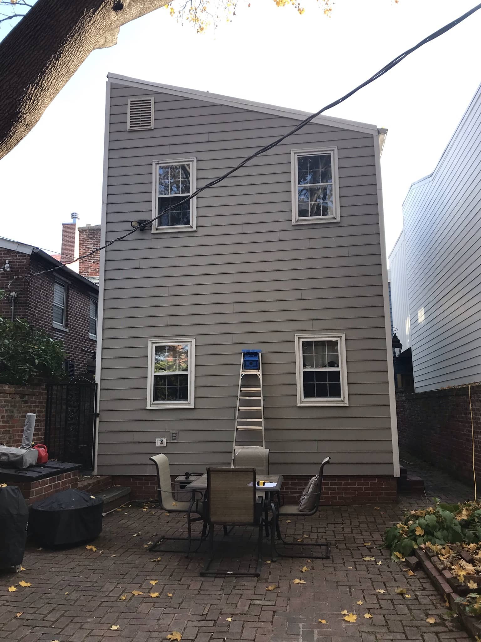 Backyard scene with a gray house featuring four windows, a ladder leaning against the wall, a table with chairs, and fallen leaves on the brick patio.