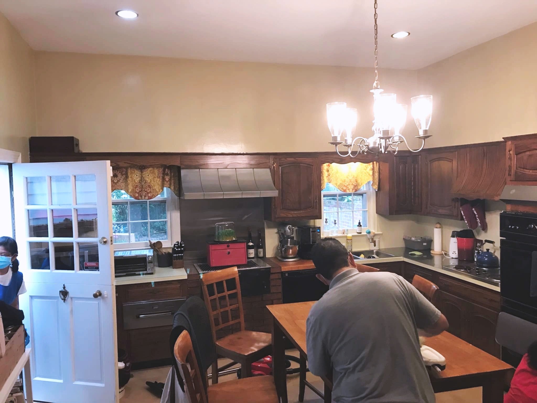 A kitchen with wooden cabinets, a dining table, and a chandelier hanging from the ceiling. Two windows with yellow and red curtains, and a person with short dark hair leaning over the table. A child wearing a face mask is partially visible on the left.