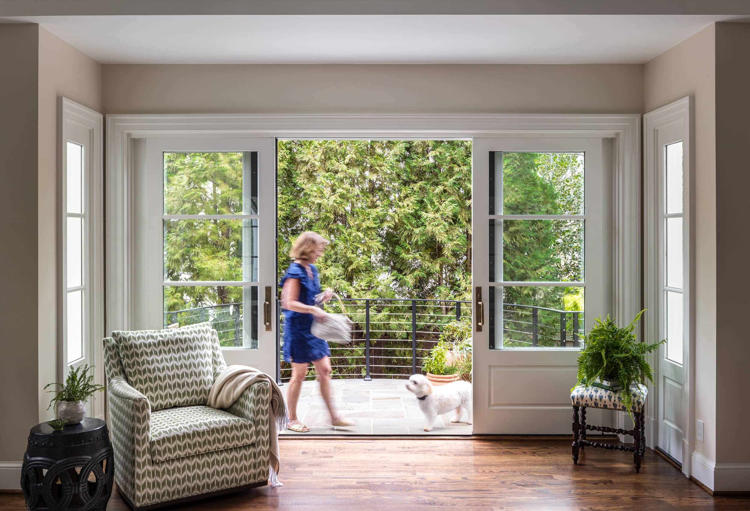Living room with open glass doors leading to a balcony, woman in blue dress walking a dog outside, green trees in background, armchair, potted plants inside.
