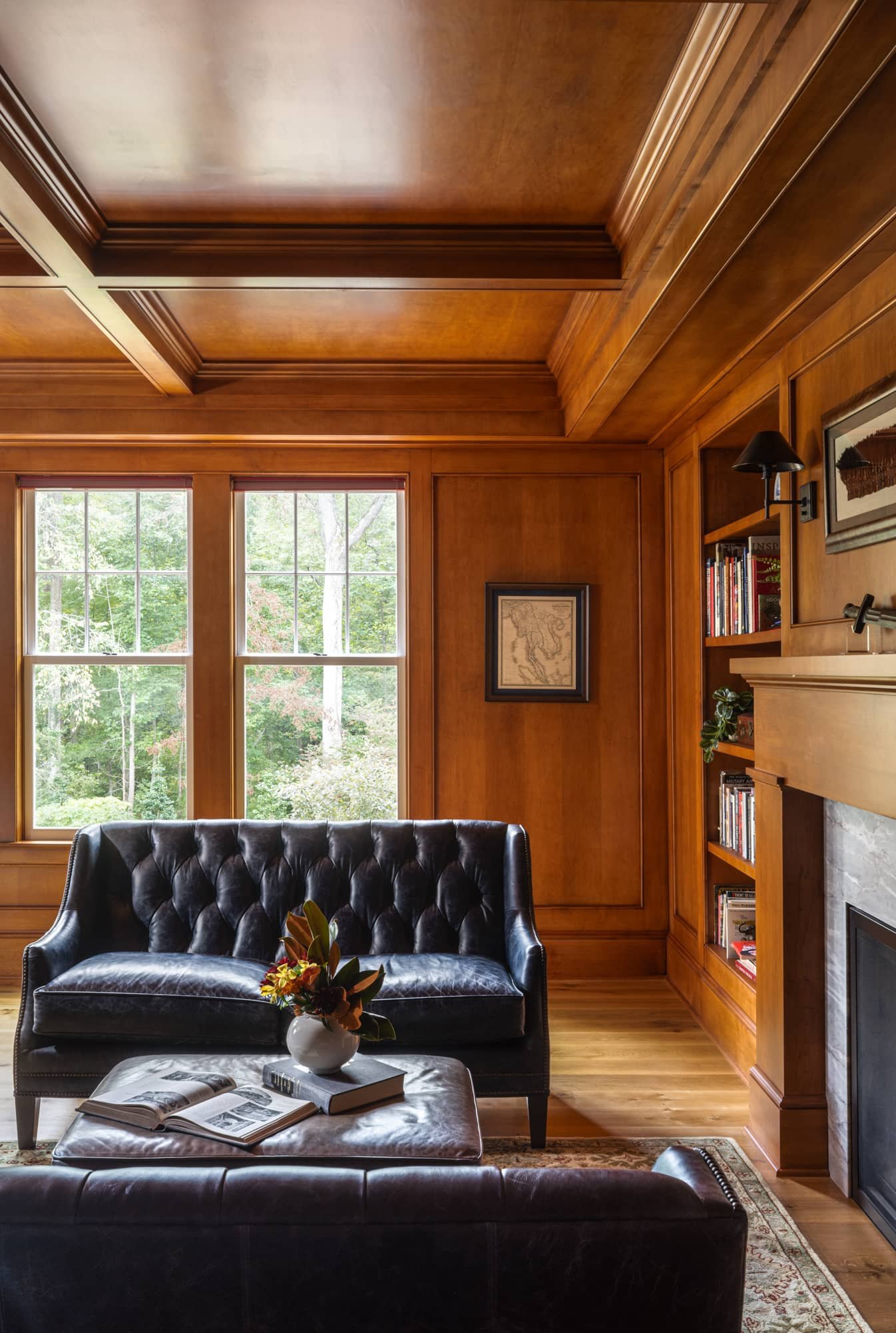 Cozy living room with large windows, wooden walls and ceiling, black leather sofa, coffee table with open book, framed pictures, bookshelf, and fireplace.