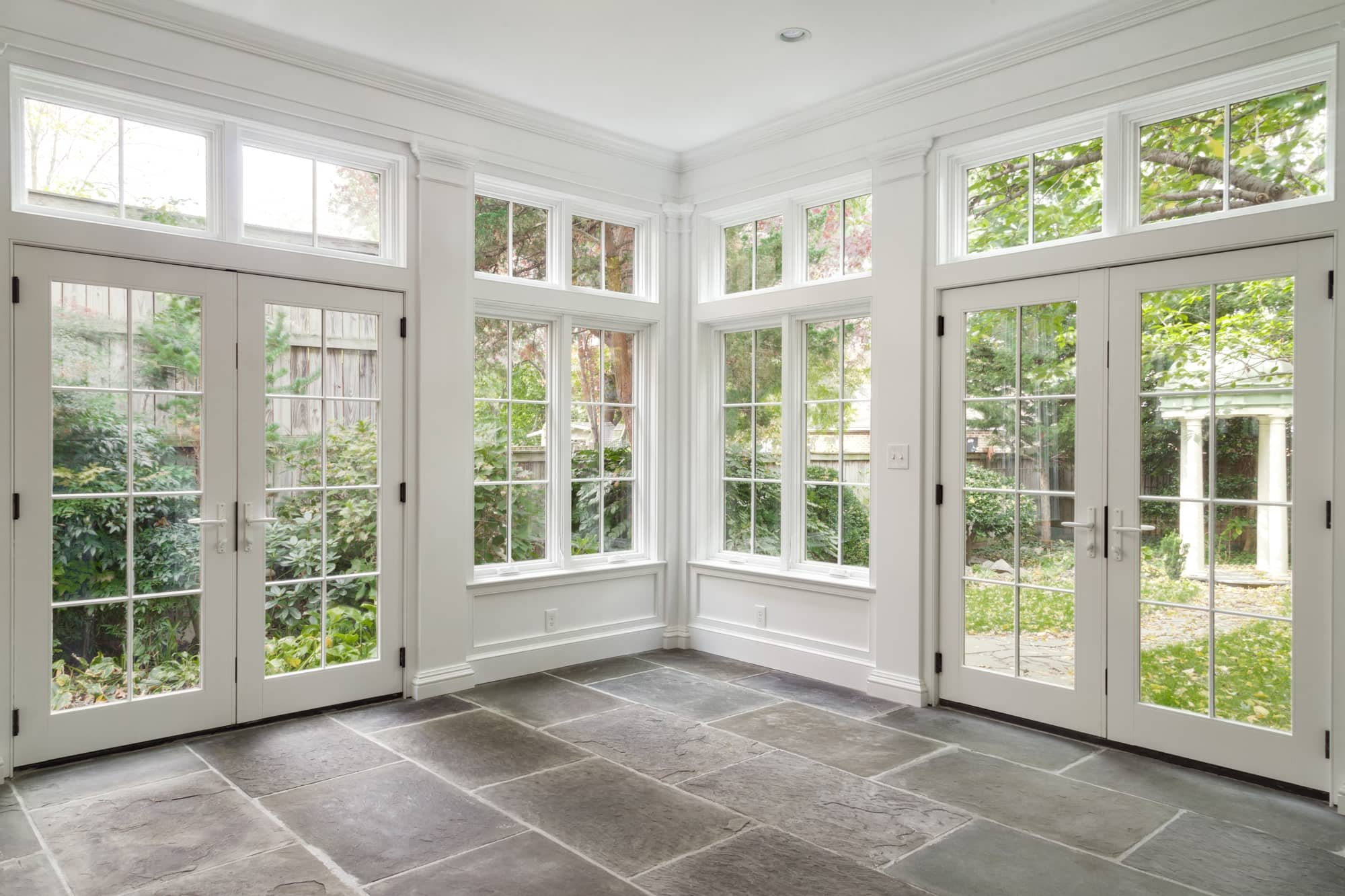 Bright enclosed patio with large white-framed glass doors and windows, stone tile flooring, and a view of a lush garden outside.