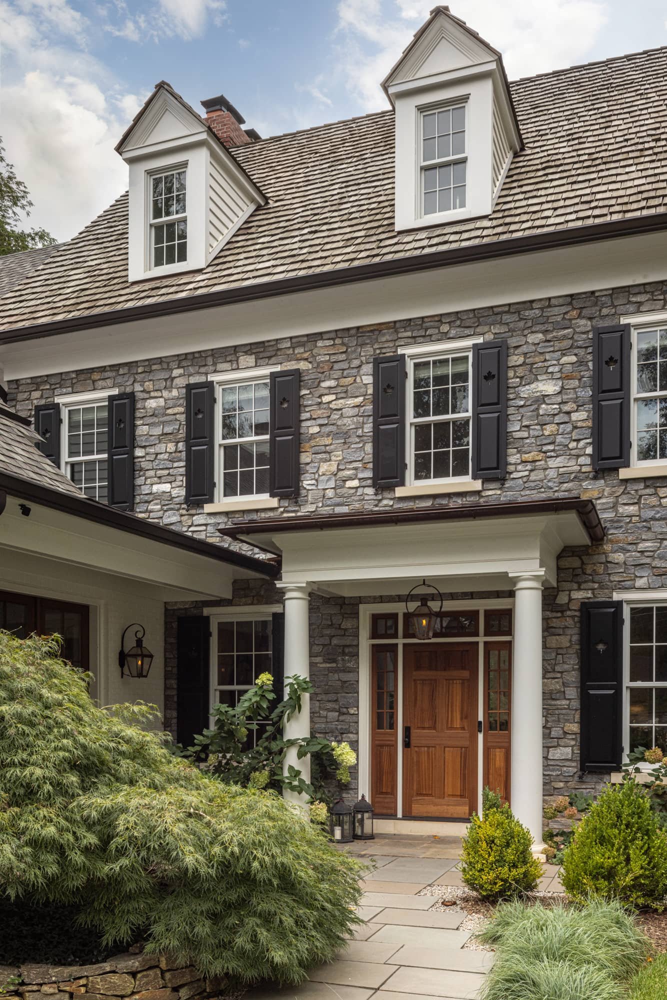 Front view of a classic Colonial house with stone exterior, black shutters, white dormer windows, and a wooden front door, surrounded by greenery and a stone pathway.