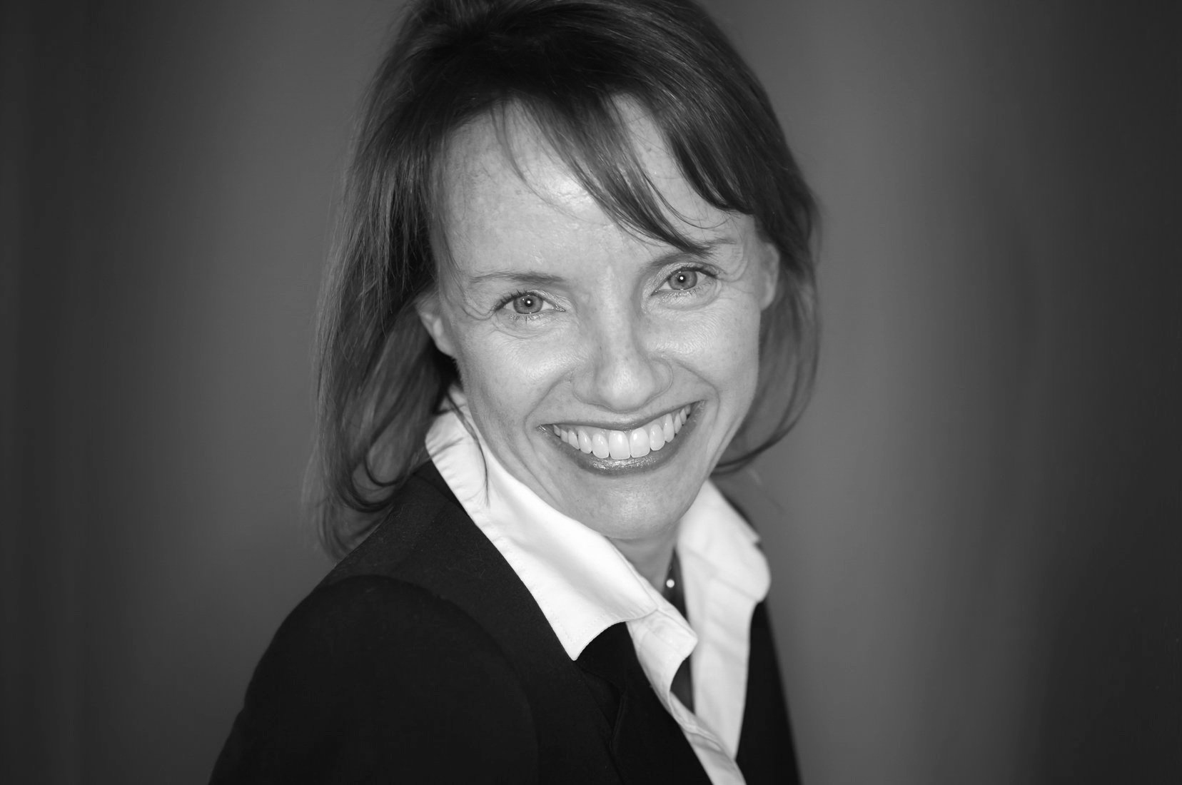 Black and white headshot of a woman with shoulder-length hair, wearing a white shirt and dark blazer, smiling at the camera.