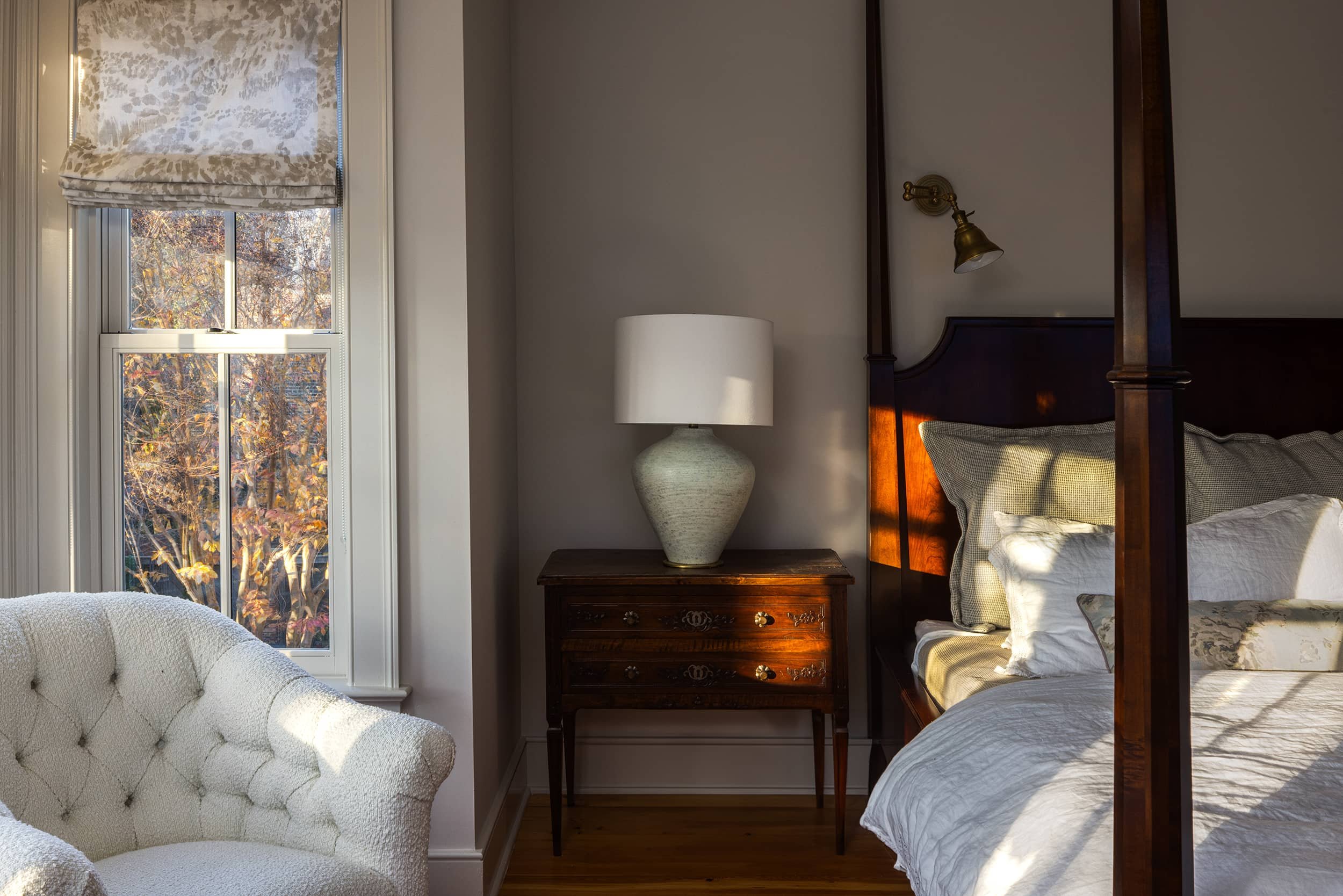 A bedroom with a bed, nightstand with lamp, window with floral shade, and a cream upholstered chair, all illuminated by natural sunlight.
