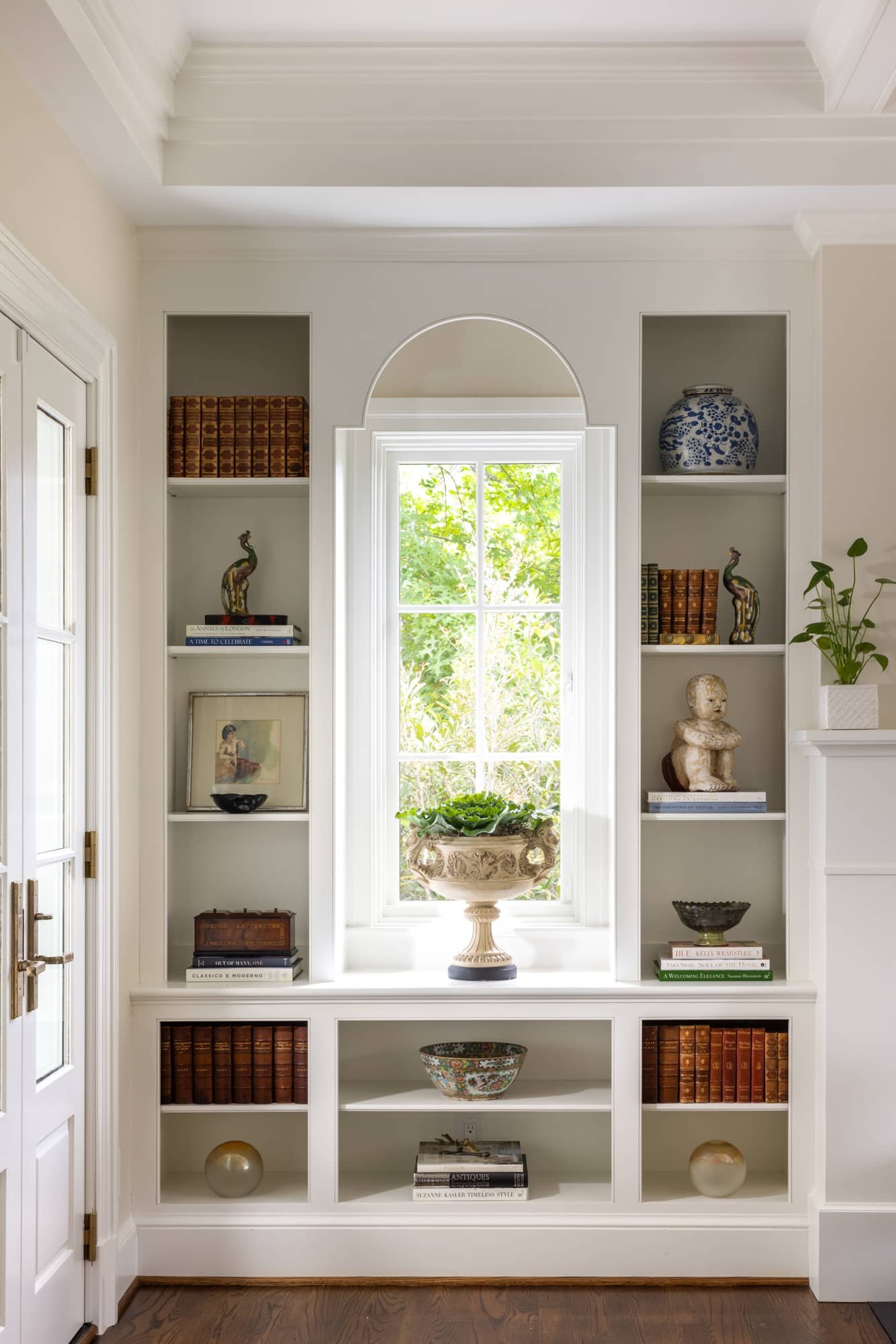 Interior view of a built-in white bookshelf with decorative items, books, and a window with green foliage outside.