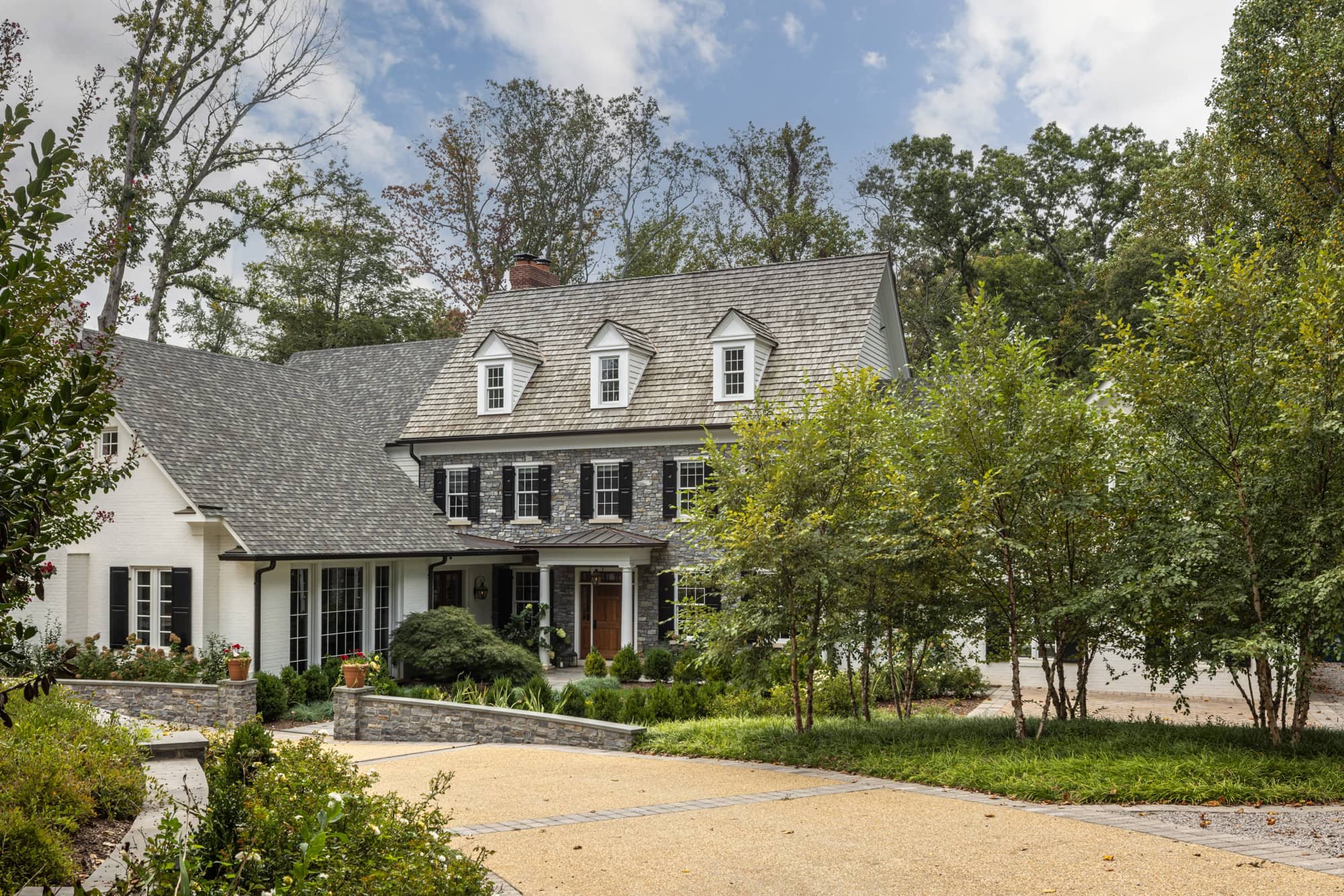 A two-story house with a gray and white exterior, black shutters, a pitched roof, and lush landscaping in front, surrounded by trees.