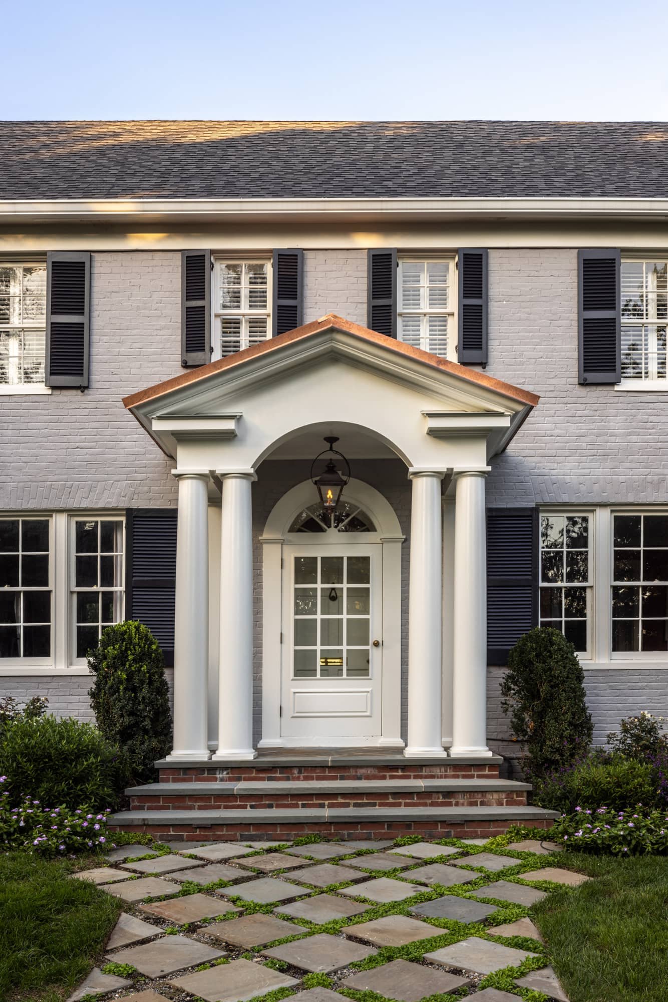 Front view of a two-story house with a brick walkway and a white porch with four pillars, black shutters, and landscaped bushes.