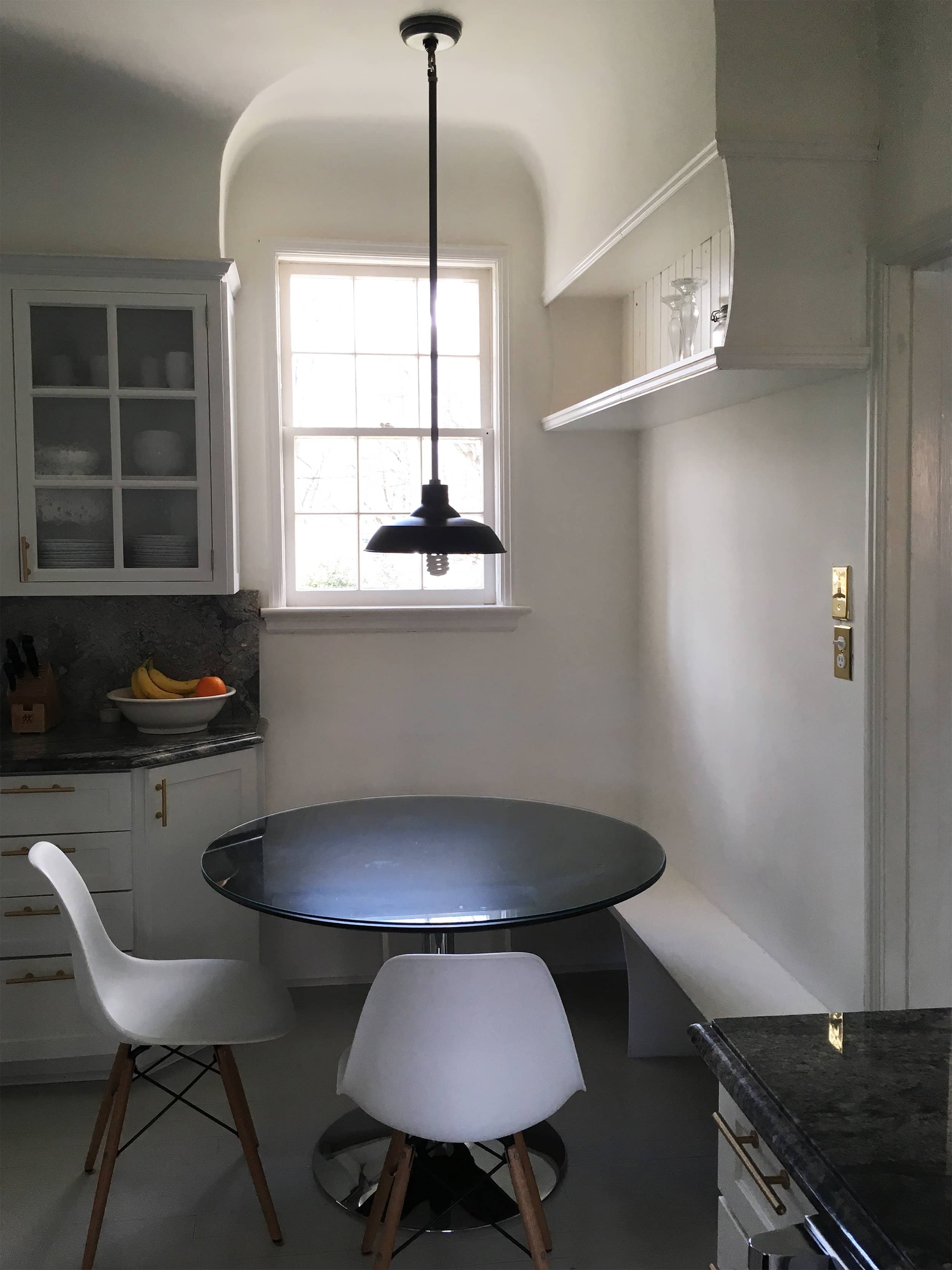 A kitchen with a round glass table, two white chairs with wooden legs, a window with sunlight shining through, and a hanging black pendant light above the table.