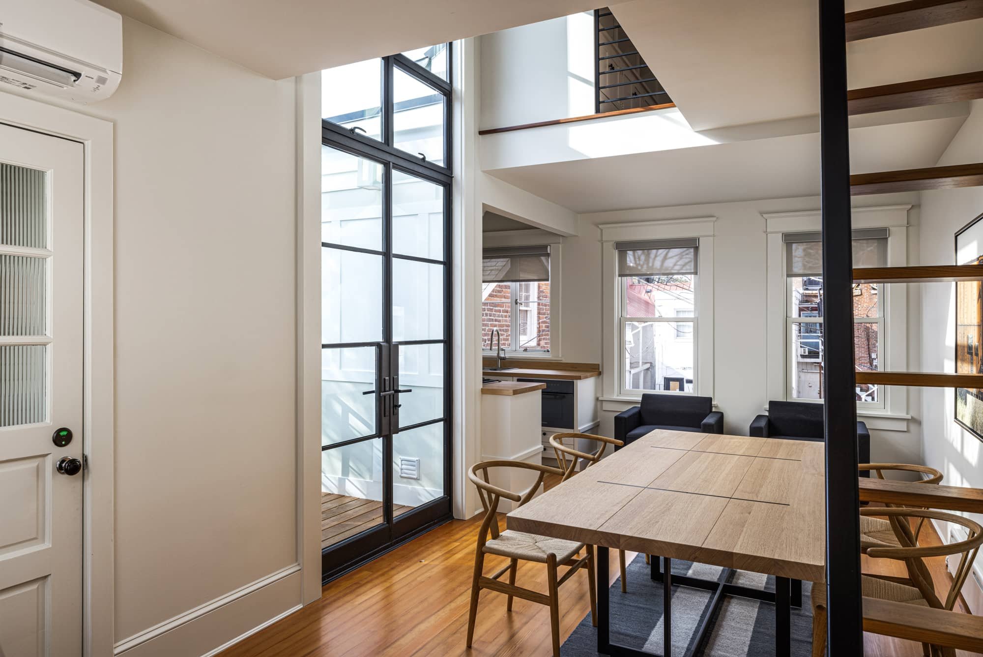 Interior of a modern, bright living space with a dining area, black chairs, a wooden table, a black armchair, windows with blinds, and a staircase with wooden steps and black supports.