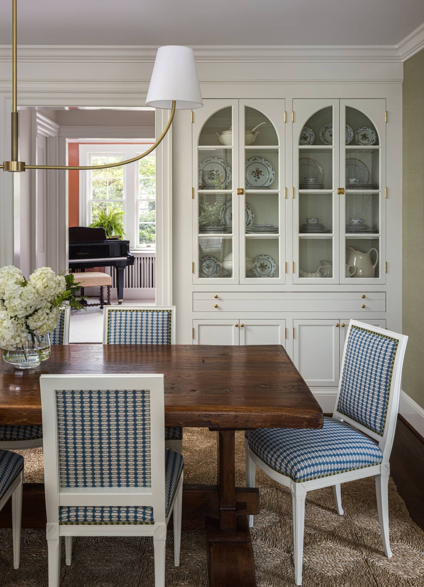 A dining room with a wooden table, four blue and white patterned chairs, a white china cabinet with dishes inside, and a vase of white flowers on the table. In the background, a living room with a window, a black piano, and a chair.