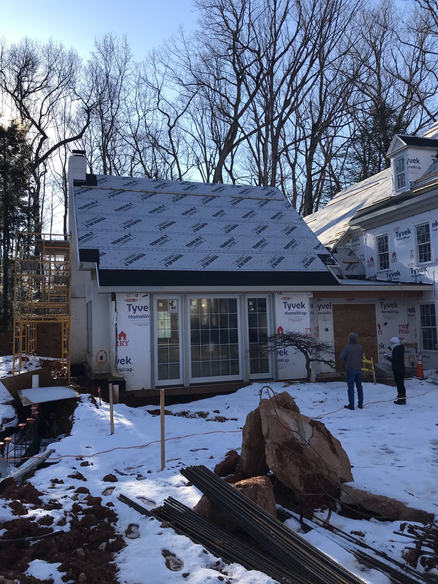 Construction site of a house during winter with snow on the ground, workers present, and new roofing being installed. The house has partially installed siding and large sliding glass doors.