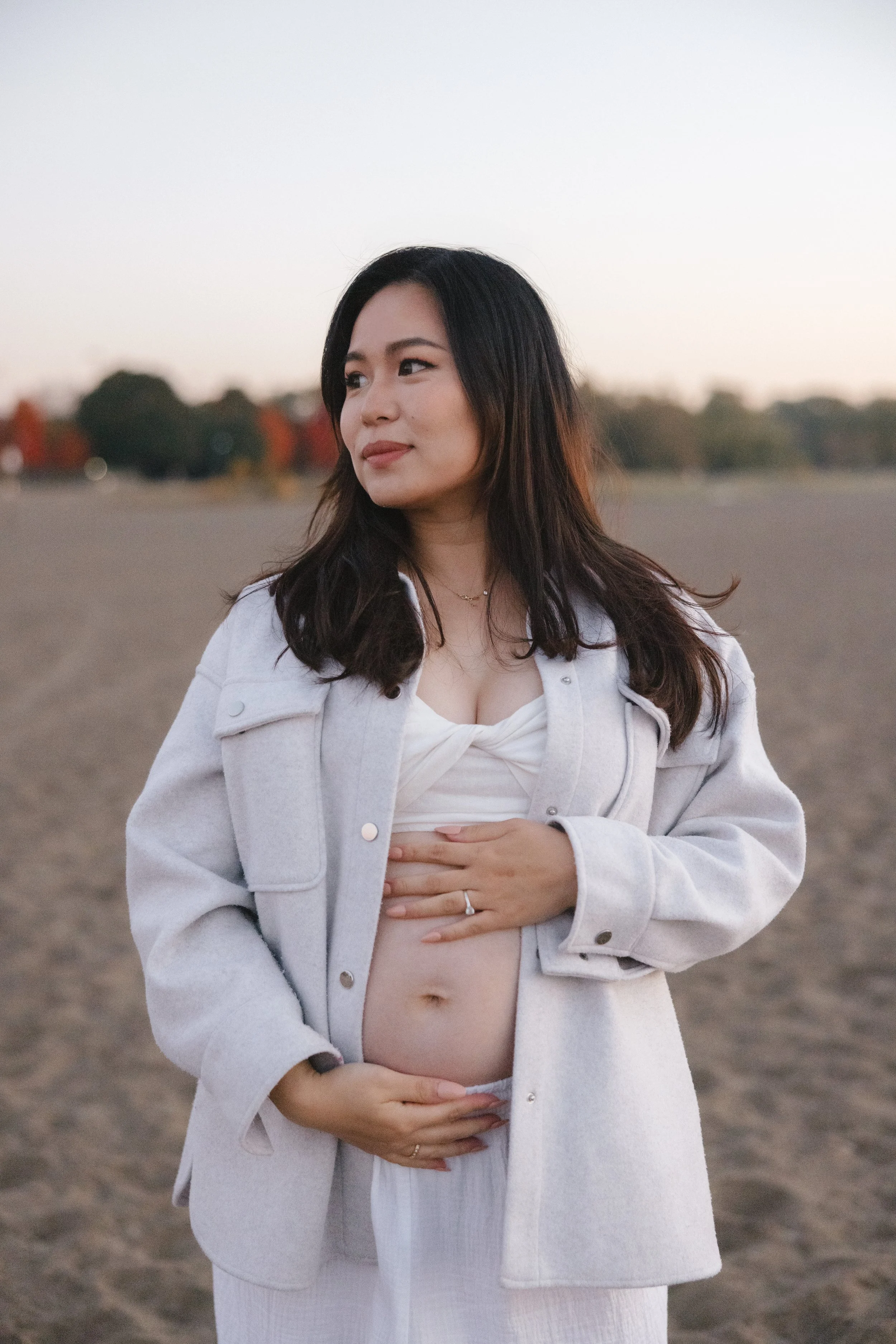 A pregnant woman with dark hair wearing a white jacket and dress, standing outdoors on a sandy area with trees in the background during sunset or dawn, holding her belly.