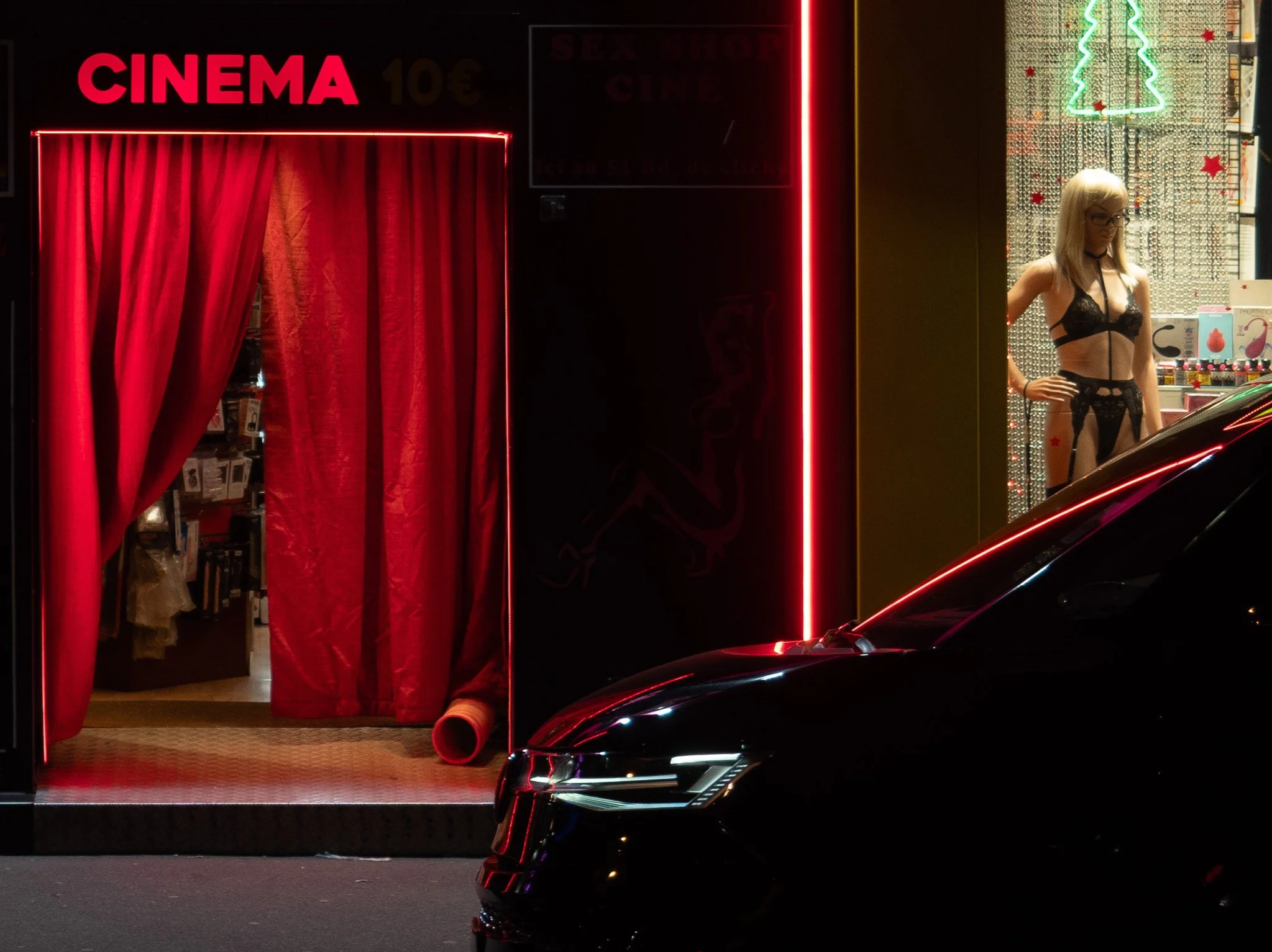 Nighttime scene of a store window display with a tag reading 'CINEMA' in red neon lights, red curtains, a woman in black lingerie, and holiday-themed decorations including a neon Christmas tree and red stars.