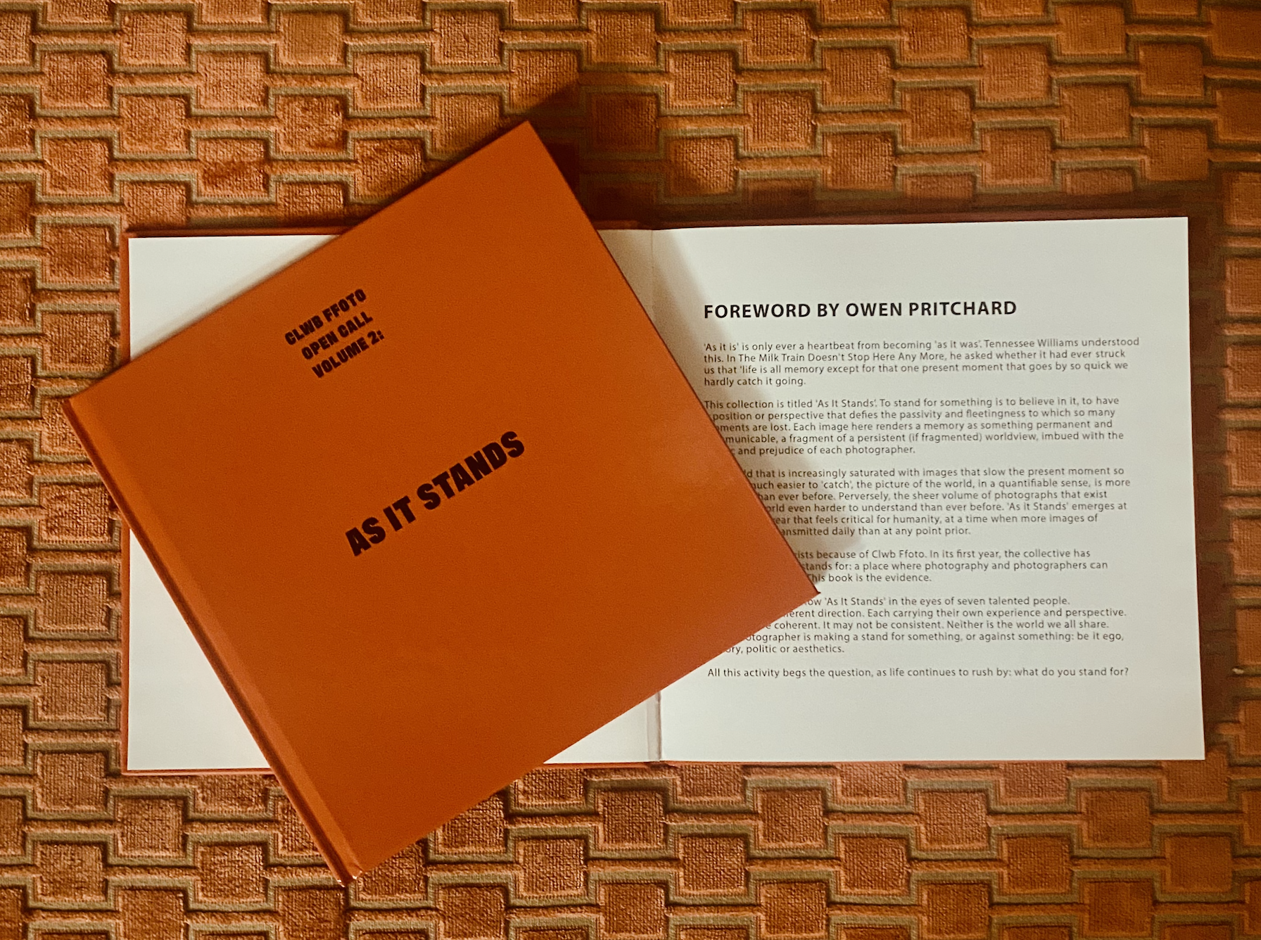 Open book titled 'AS IT STANDS' resting on top of a white sheet of paper on a textured orange surface.