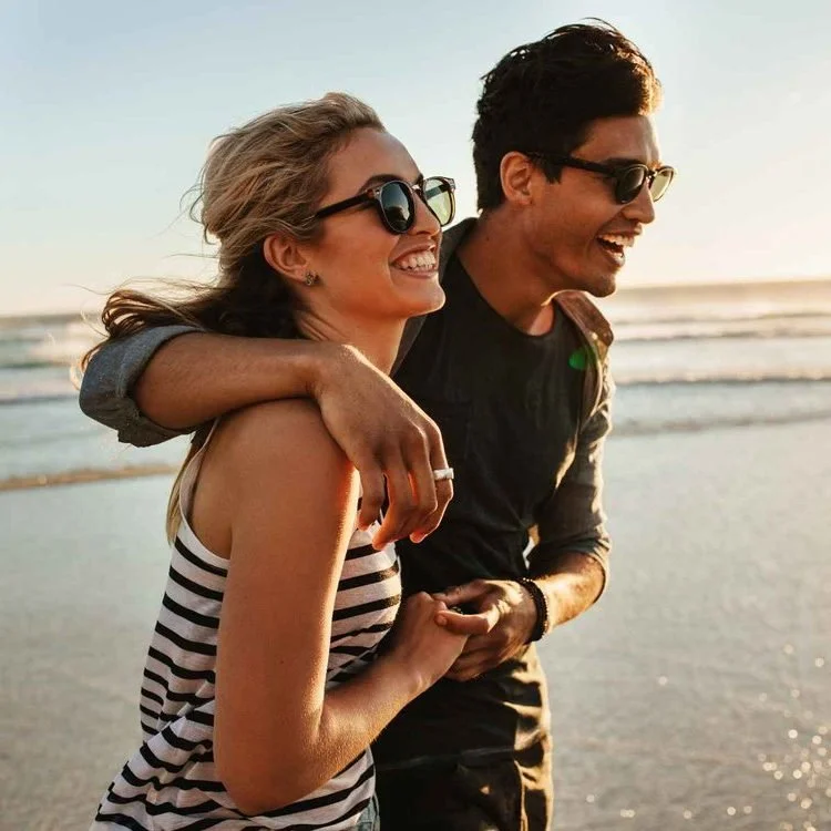 A young couple enjoying a walk on the beach at sunset, smiling and laughing, with the ocean in the background.