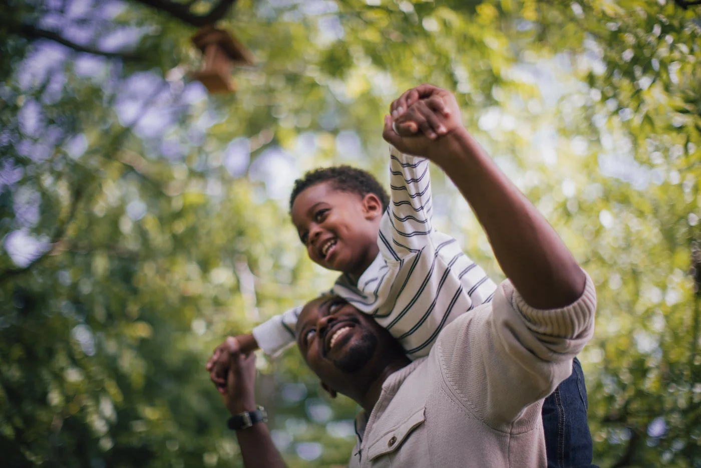 A man lifting a young boy on his shoulders outdoors among green trees, both smiling and enjoying a playful moment.