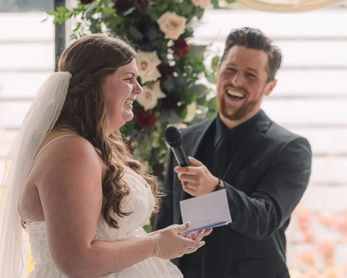A bride and groom sharing a laugh during their wedding ceremony, with the officiant holding a microphone and a notebook, in front of a floral backdrop.