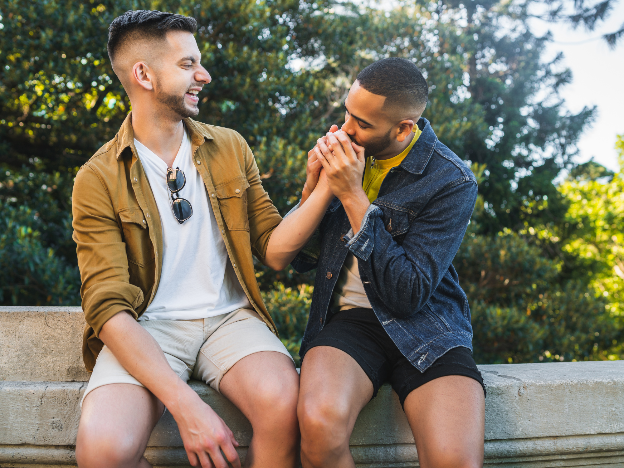 A couple sitting on a concrete bench outdoors, one smiling and touching the other's face, while the other kisses his hand, with trees and sunlight in the background.