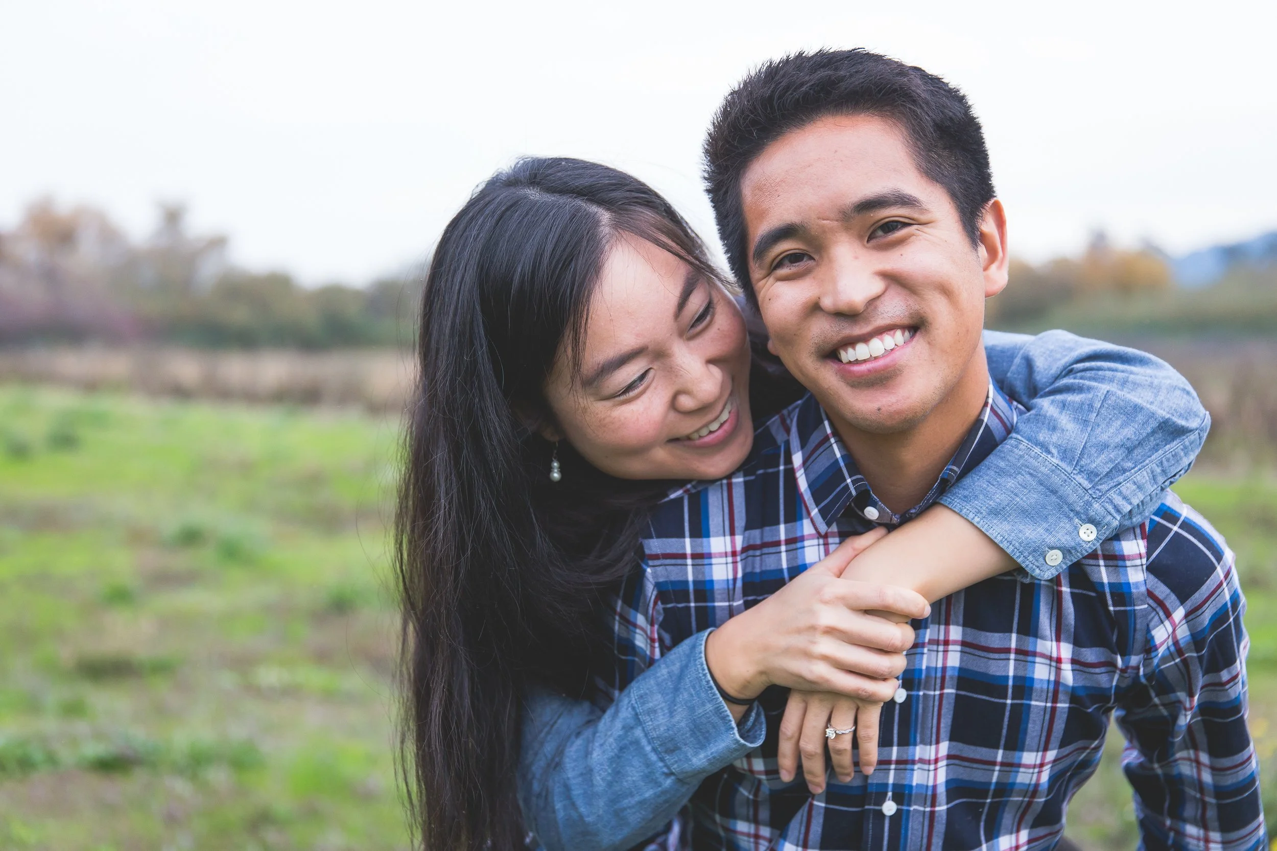 A young couple smiling outdoors, the woman hugging the man from behind.
