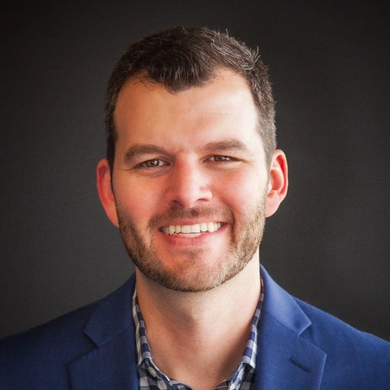 Headshot of a smiling man with short dark hair and a beard, wearing a blue blazer and checkered shirt against a dark background.
