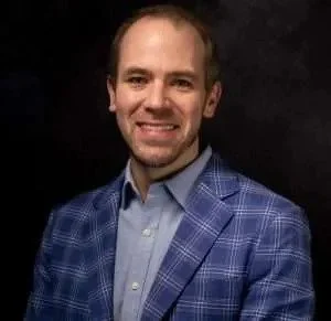 A man with short hair and a beard wearing a blue checkered blazer and light blue shirt smiling against a dark background.