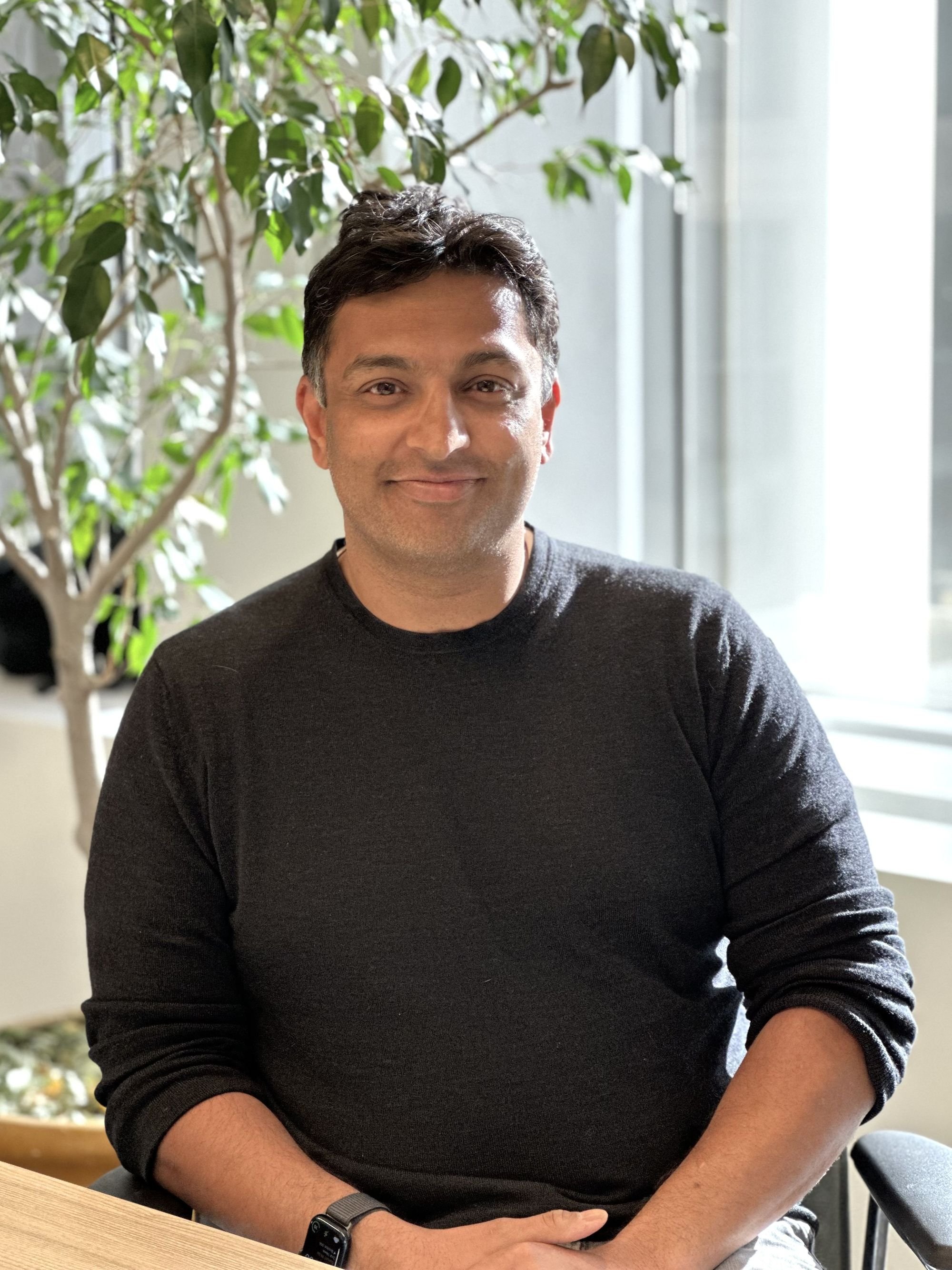 A man sitting at a table indoors, smiling, wearing a black sweater and a smartwatch, with a large green plant and window in the background.