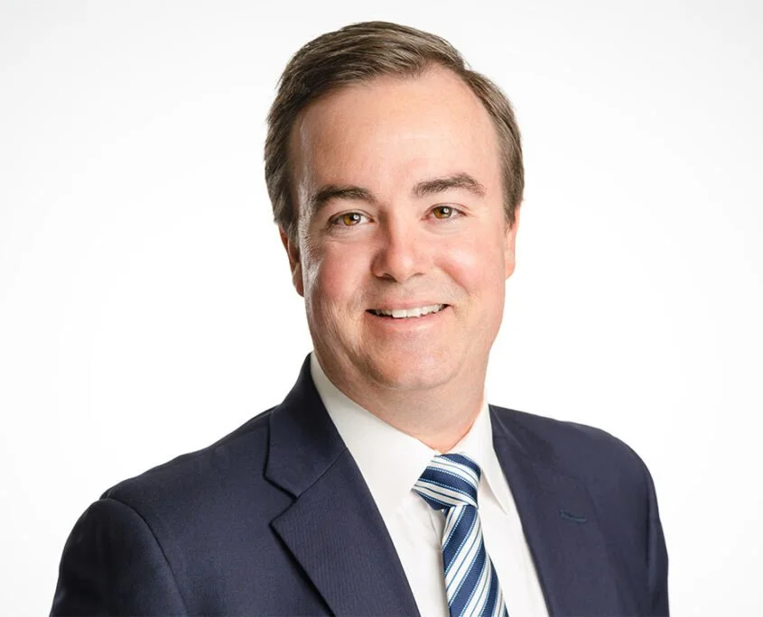 Professional headshot of a smiling man in a dark suit, white shirt, and striped tie, against a plain white background.