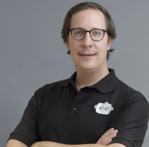 A man with glasses and brown hair, wearing a black polo shirt with a camera logo, standing with arms crossed against a gray background.