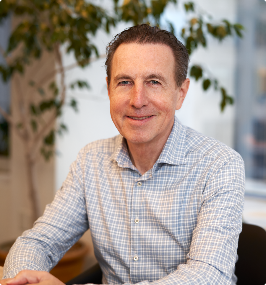 Smiling man in checkered shirt sitting at a table indoors with a plant and windows in the background.