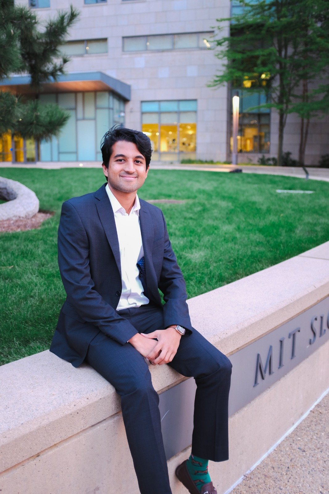 A young man dressed in a dark suit sitting on a stone ledge outside of MIT, smiling, with a modern building and green trees in the background.